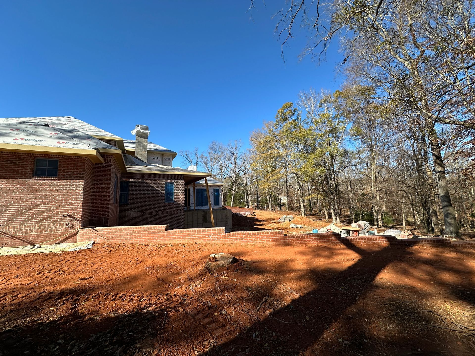 Red brick building under construction with exposed red soil, and trees under a clear blue sky.
