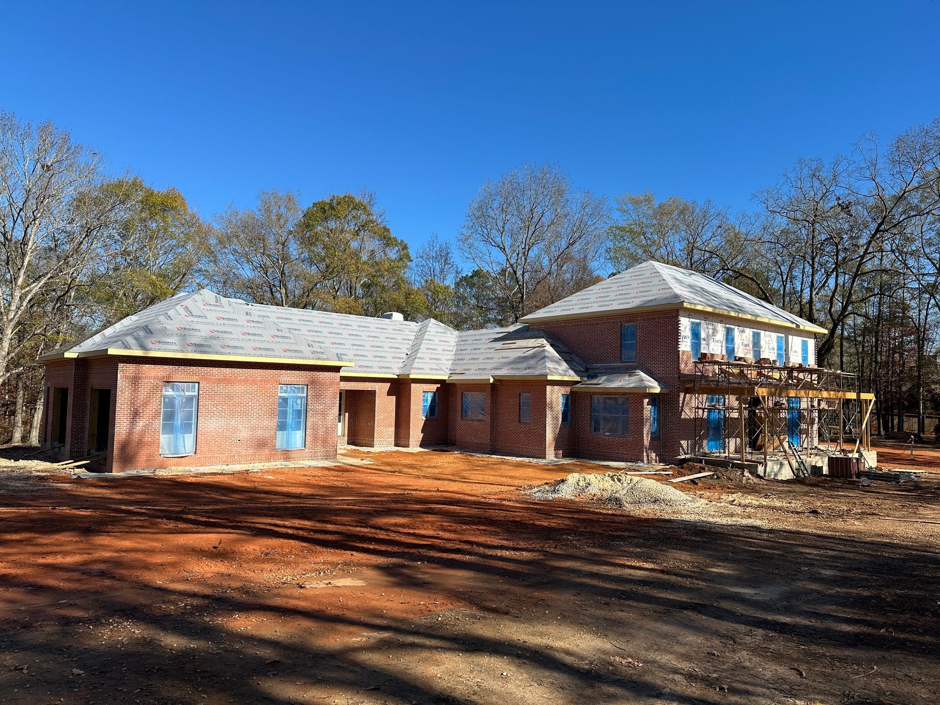 New brick house under construction on a dirt lot, with a blue sky and bare trees in the background.