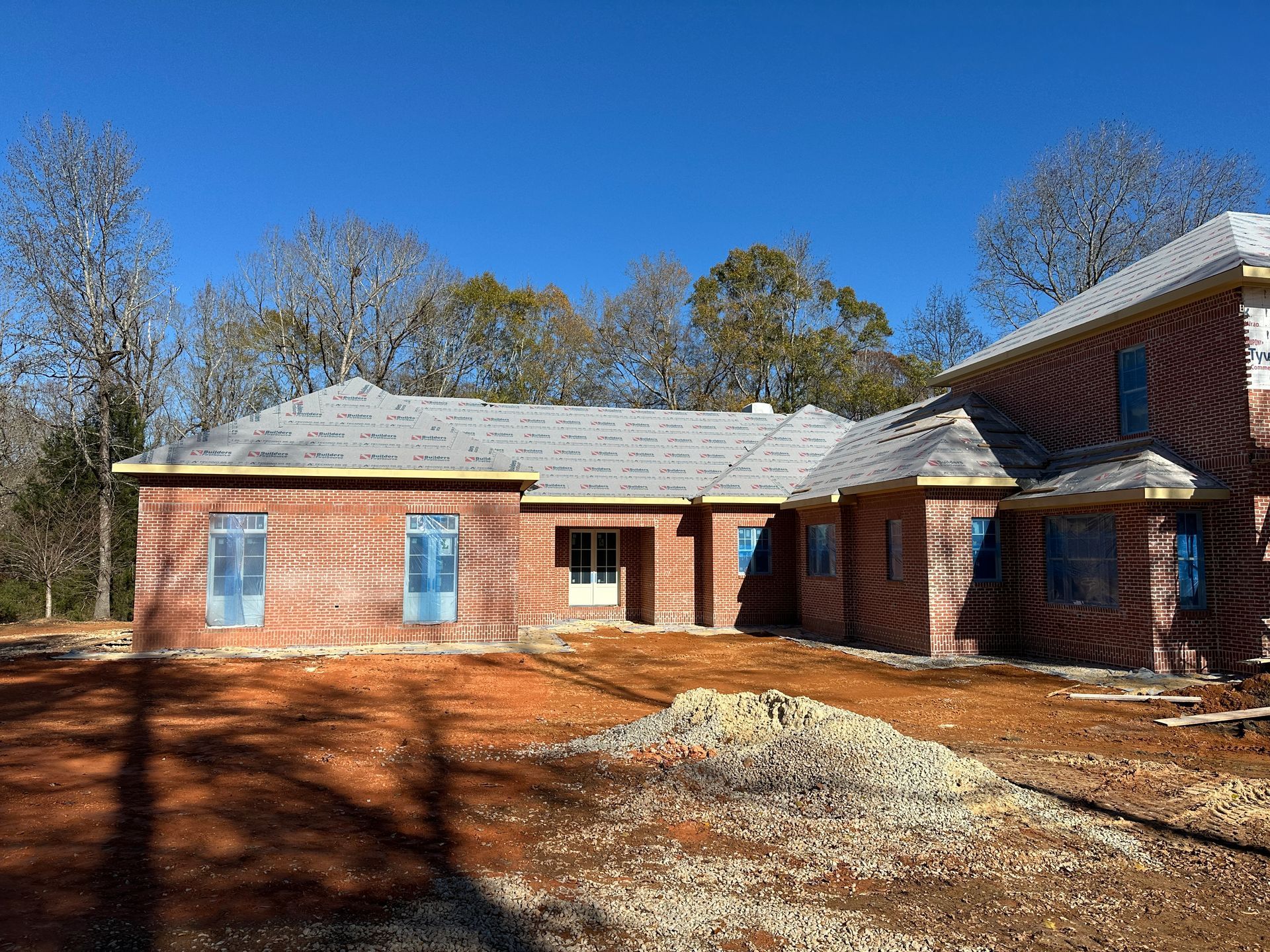 Brick house under construction with a gray roof and blue-covered windows in a yard.