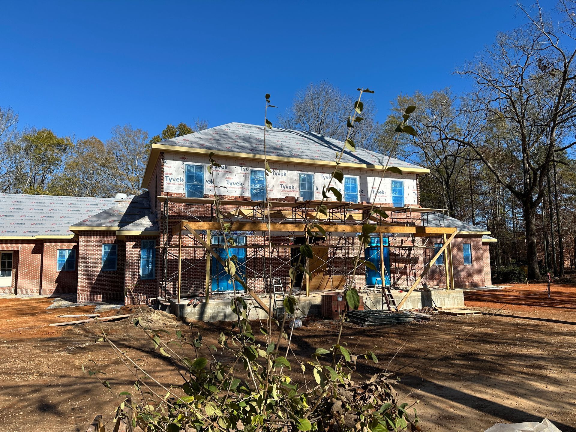 Two-story house under construction with exposed wood framing, blue coverings, and surrounding trees against a blue sky.