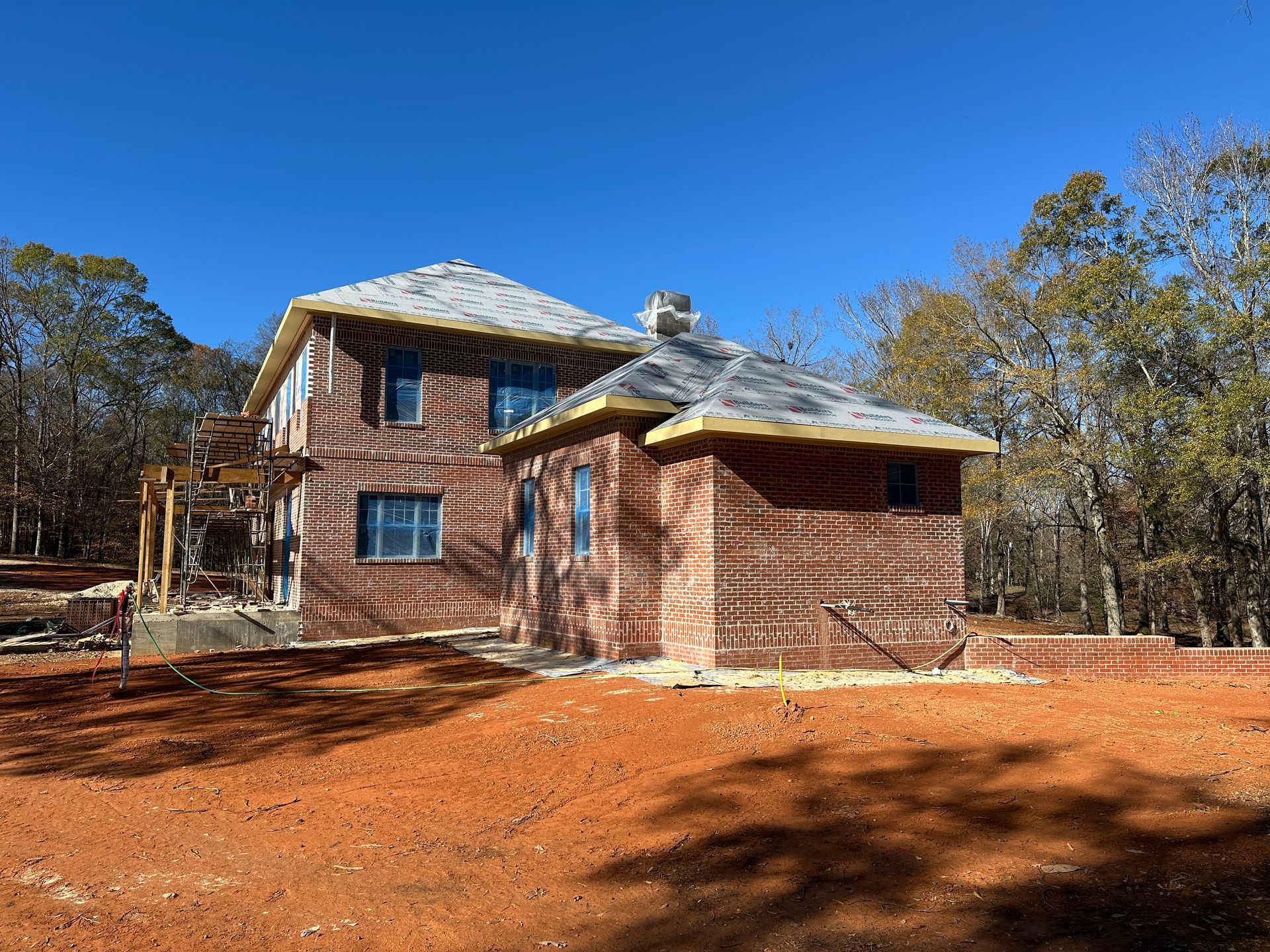 Two-story brick house under construction, surrounded by trees and reddish-brown soil, clear blue sky.