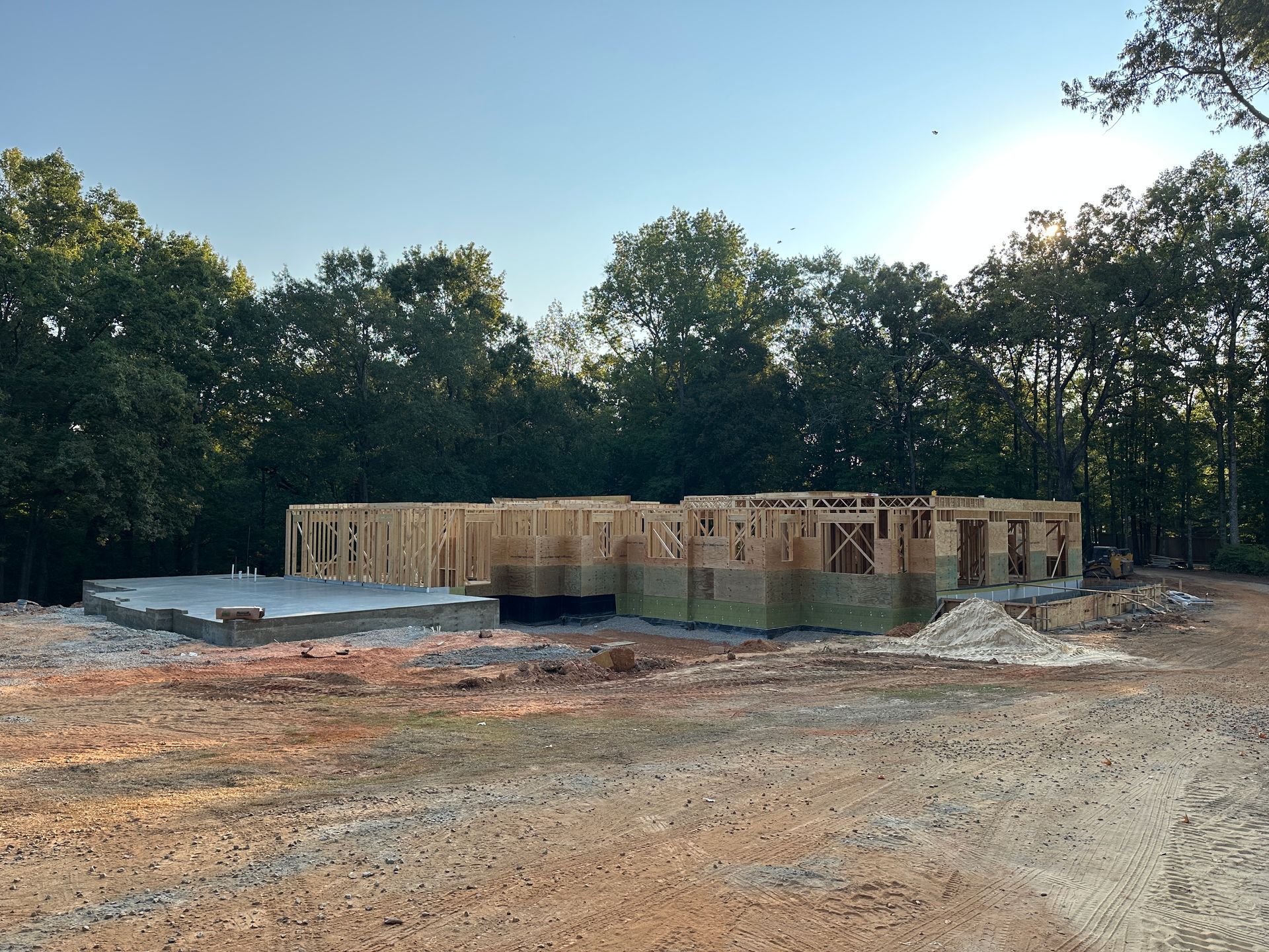 Construction site: wooden frame of a house on a concrete foundation, surrounded by dirt and trees.