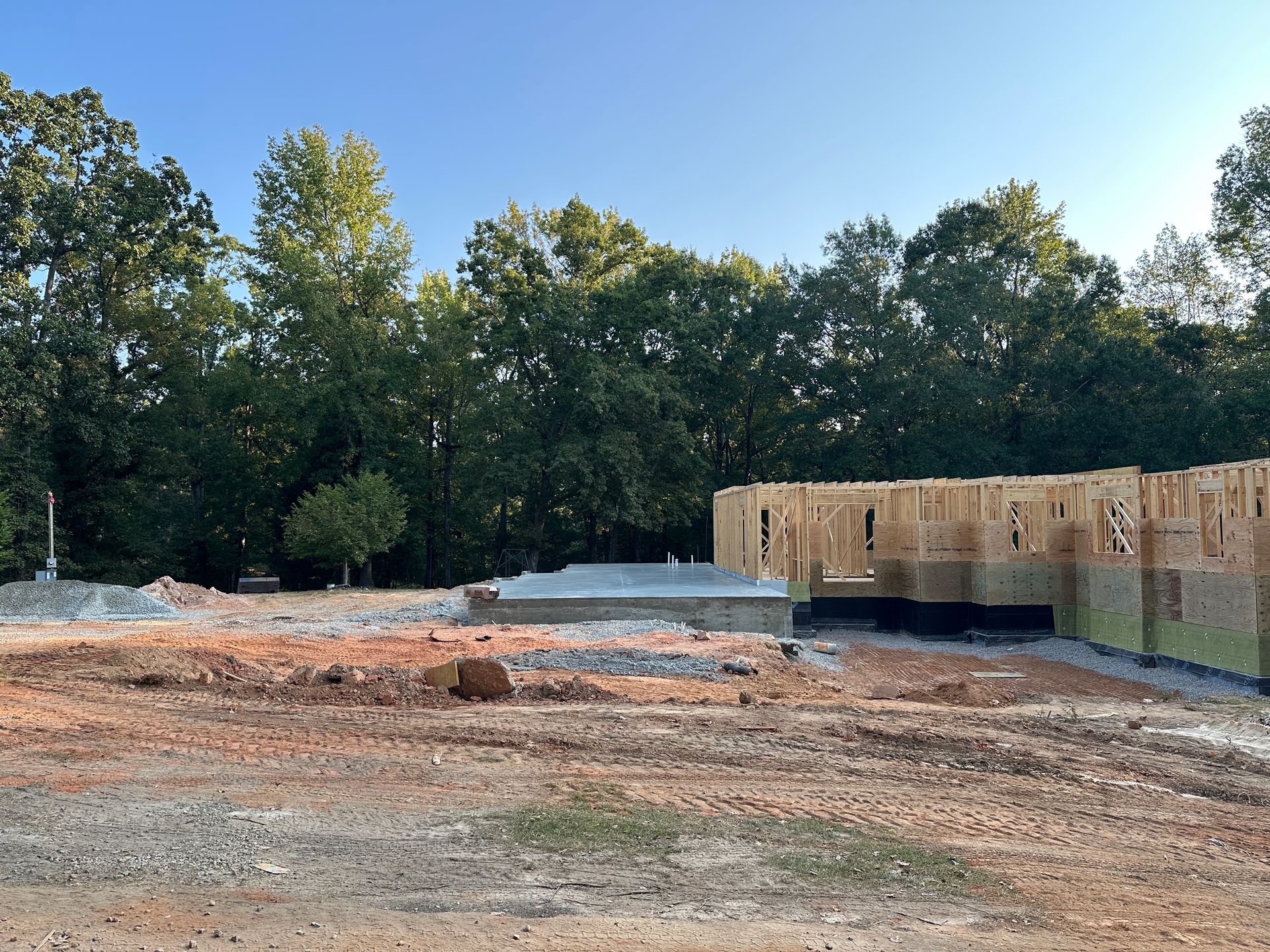 Construction site with wood framing, concrete foundation, and trees under a blue sky.