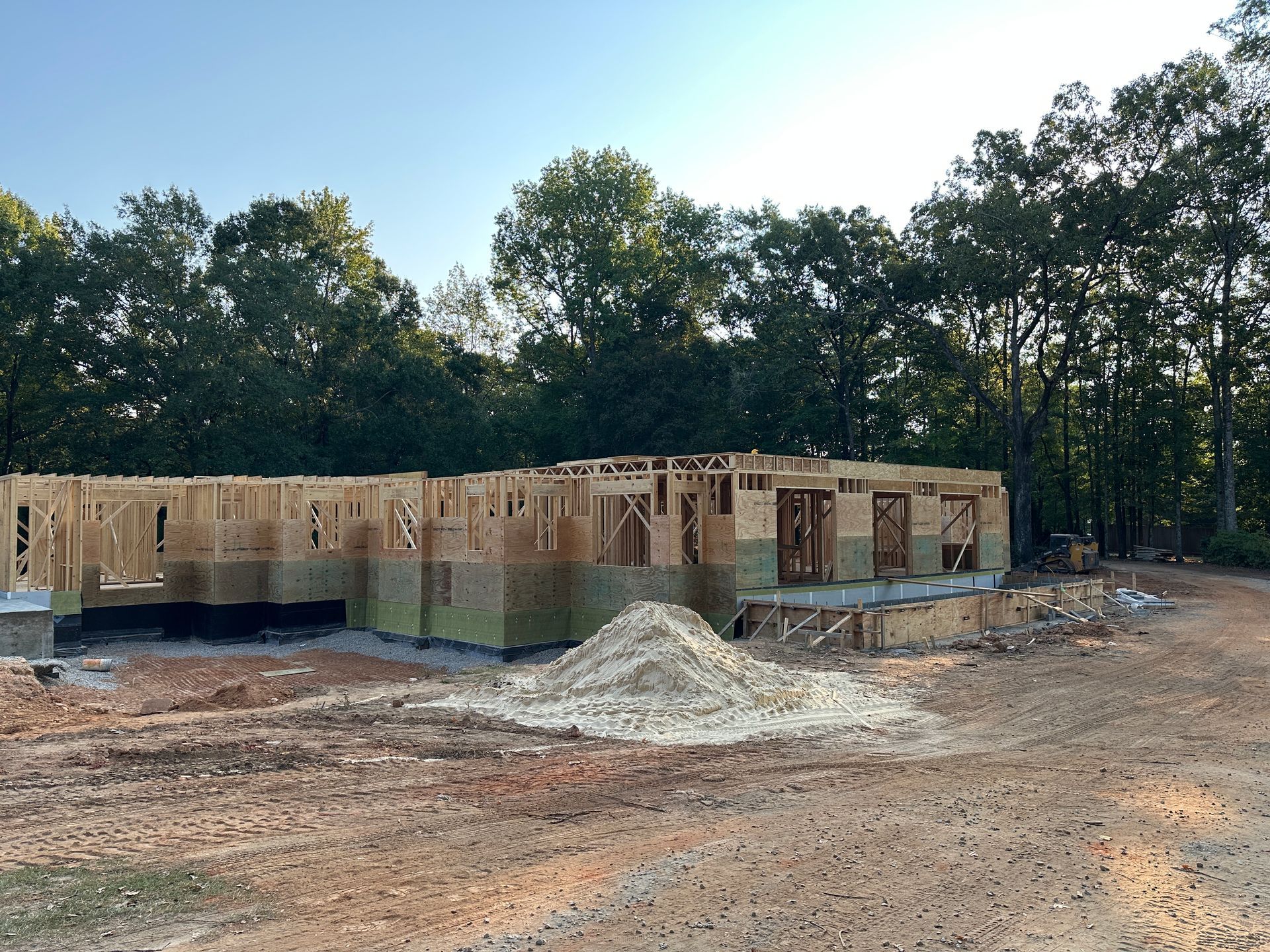 Framed wooden structure of a house under construction, surrounded by dirt and trees, under a clear sky.