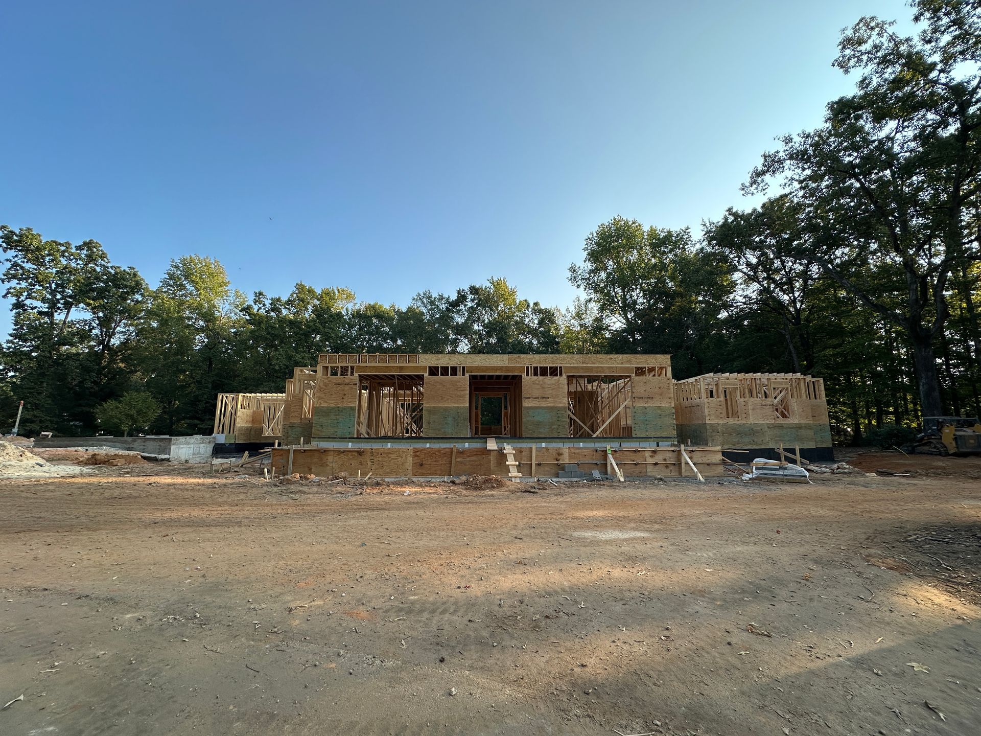 Construction site: unfinished wood-frame house against a blue sky, surrounded by trees, dirt lot in foreground.
