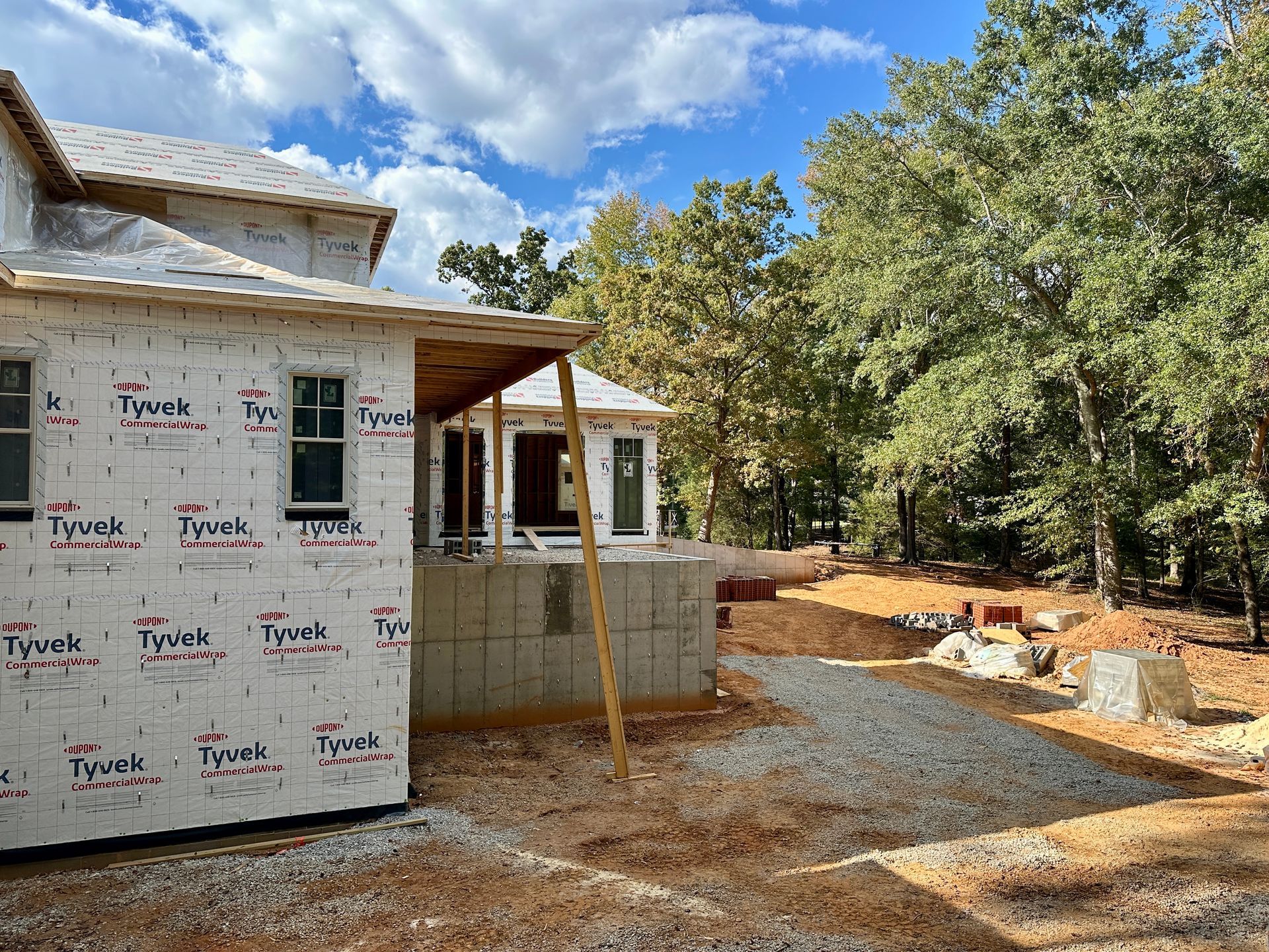Construction site with house frame covered in Tyvek wrap, gravel driveway, and surrounding trees.