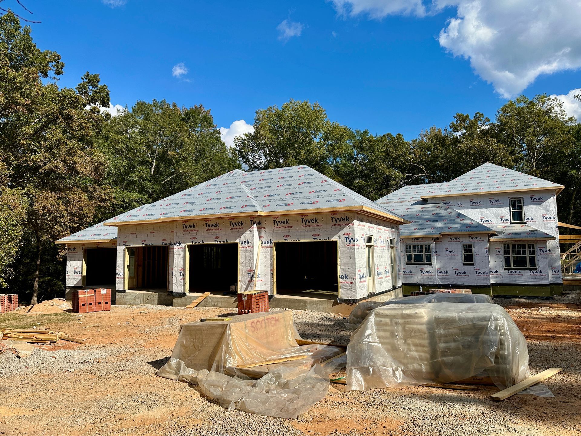 House under construction with exposed framing, blue sky, and surrounding trees.