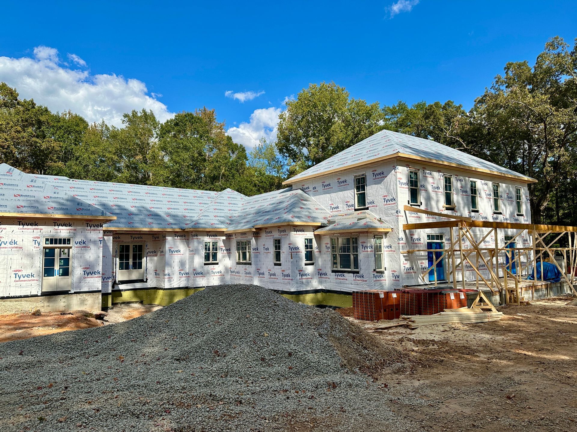 House under construction, exterior view. Blue sky, white wrap, gray gravel pile. Wood framing visible.