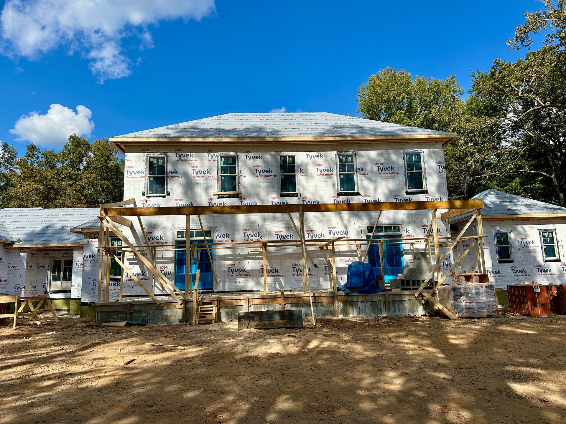 Building under construction with white protective wrap and blue doors, set against a blue sky with trees.