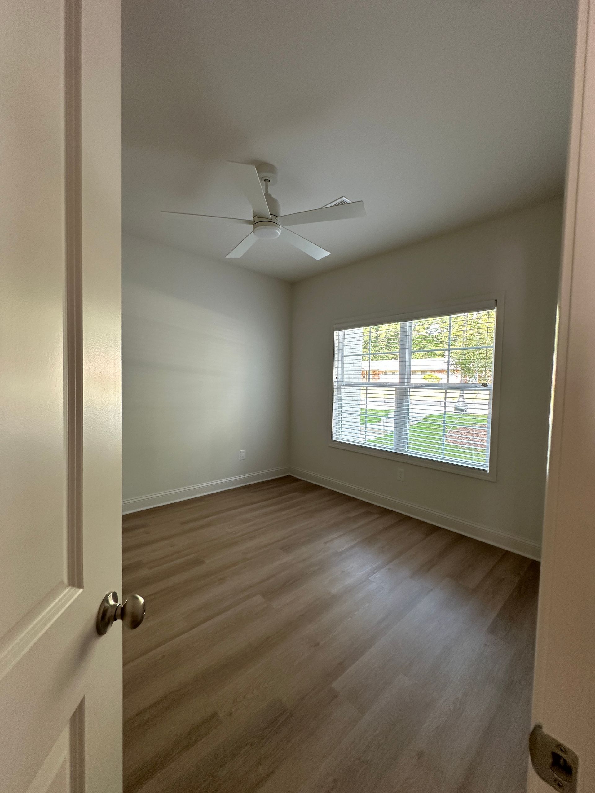 Empty bedroom with wood floors, white walls, ceiling fan, and a window with blinds.