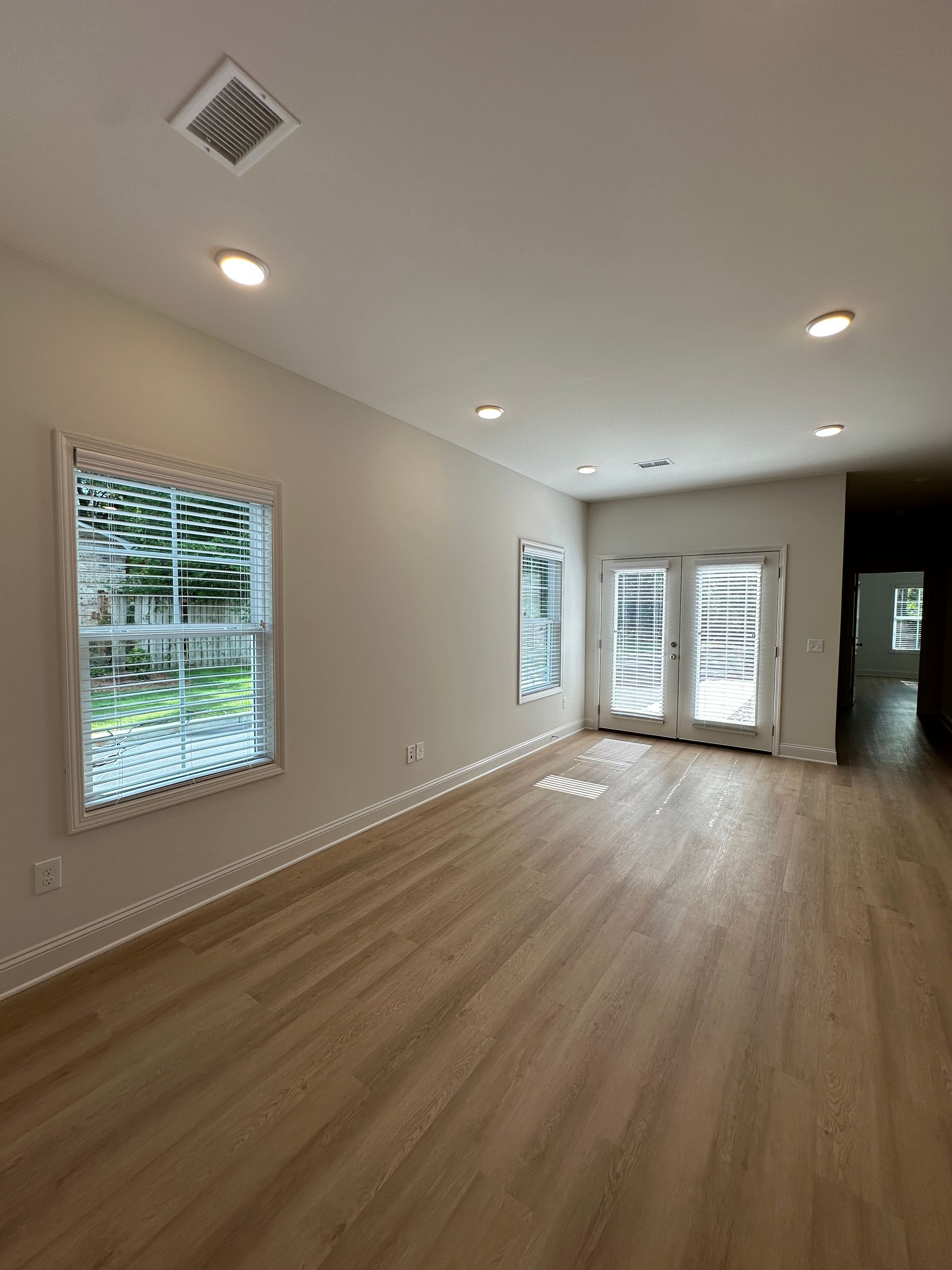 Empty modern living room with light wood floors, recessed lights, and large windows and French doors.