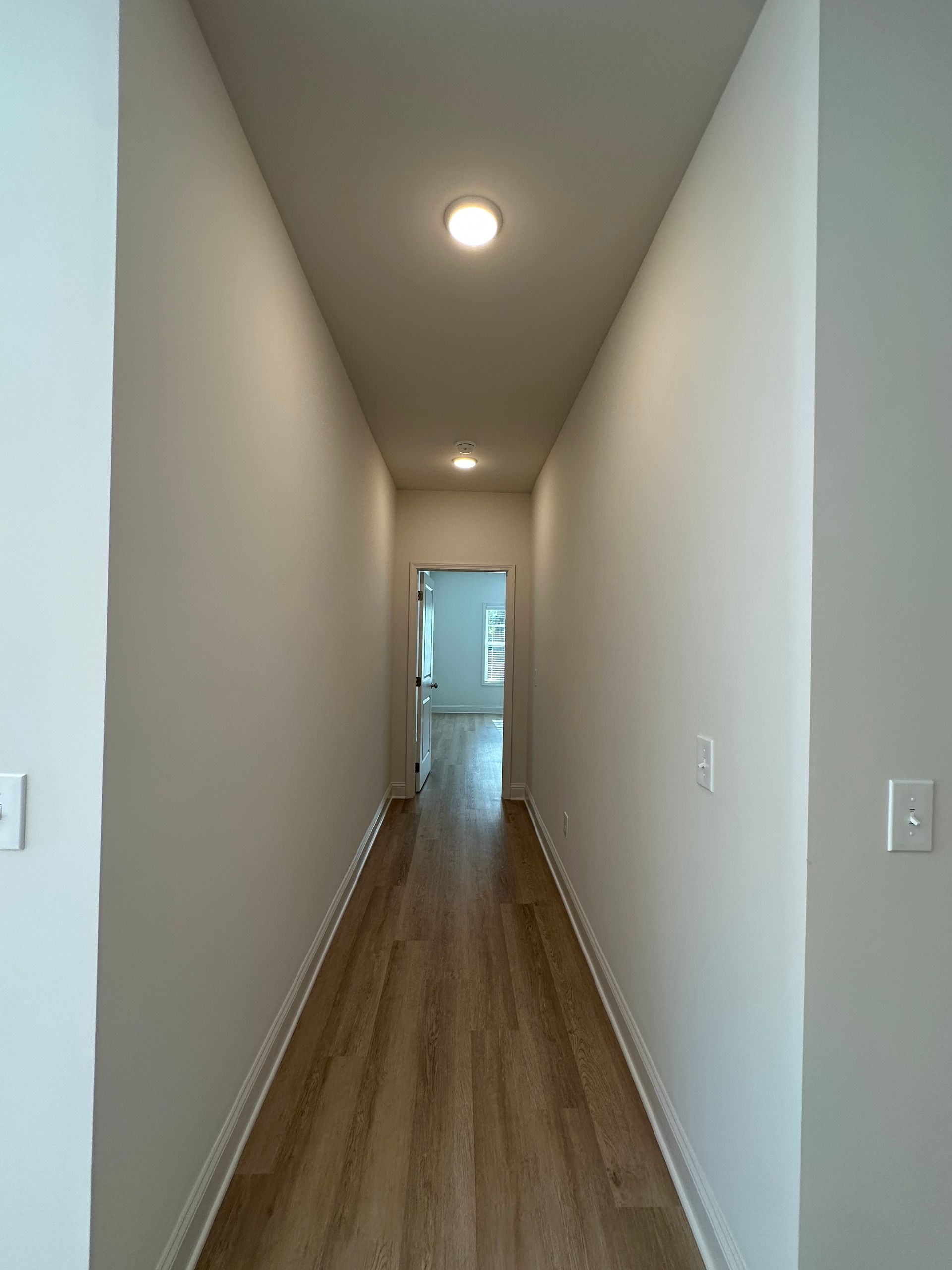 Narrow hallway with wood floors, beige walls, and recessed ceiling lights leading to a doorway.
