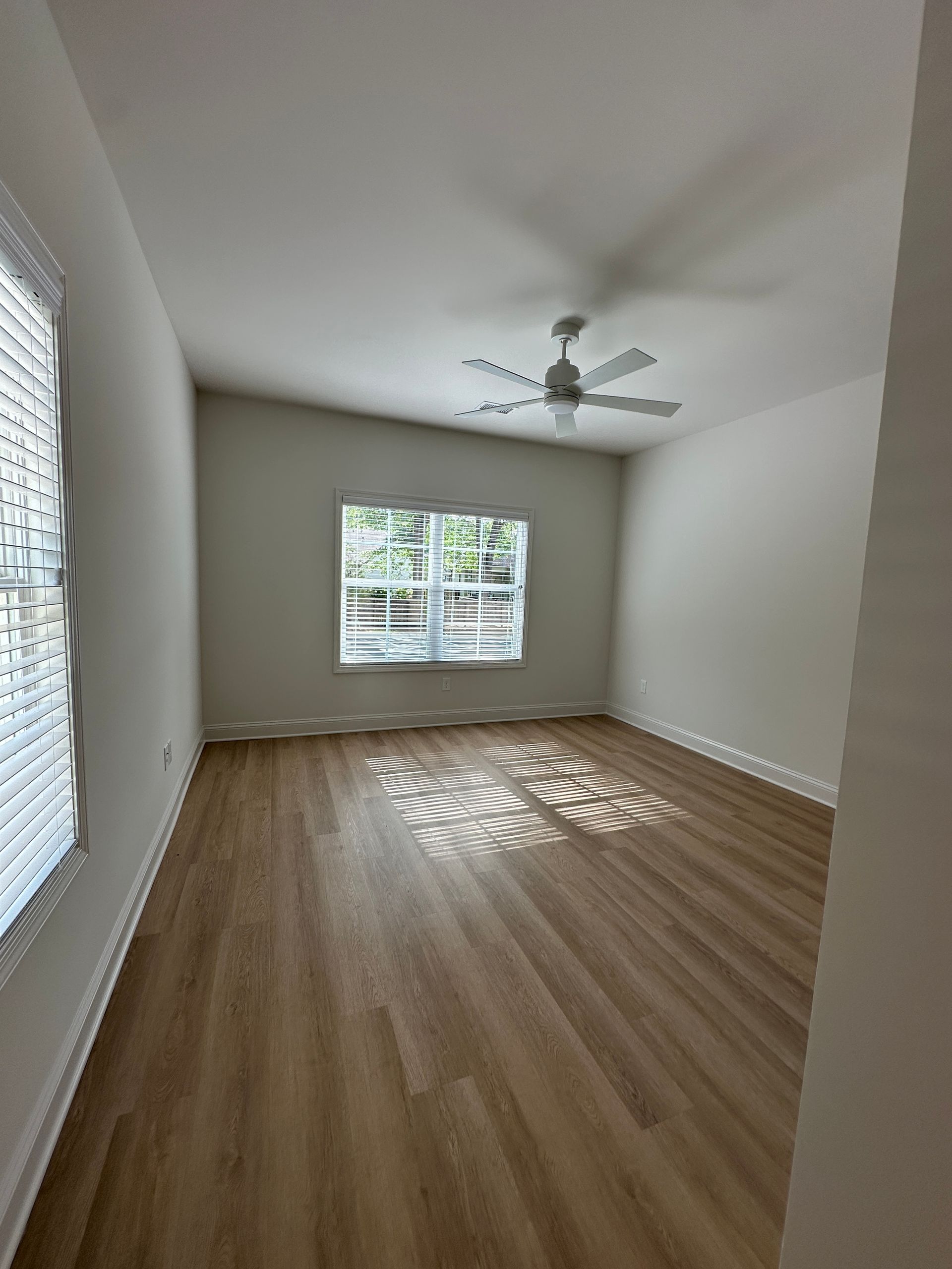 Empty beige bedroom with wood floors, ceiling fan, and sunlight through windows