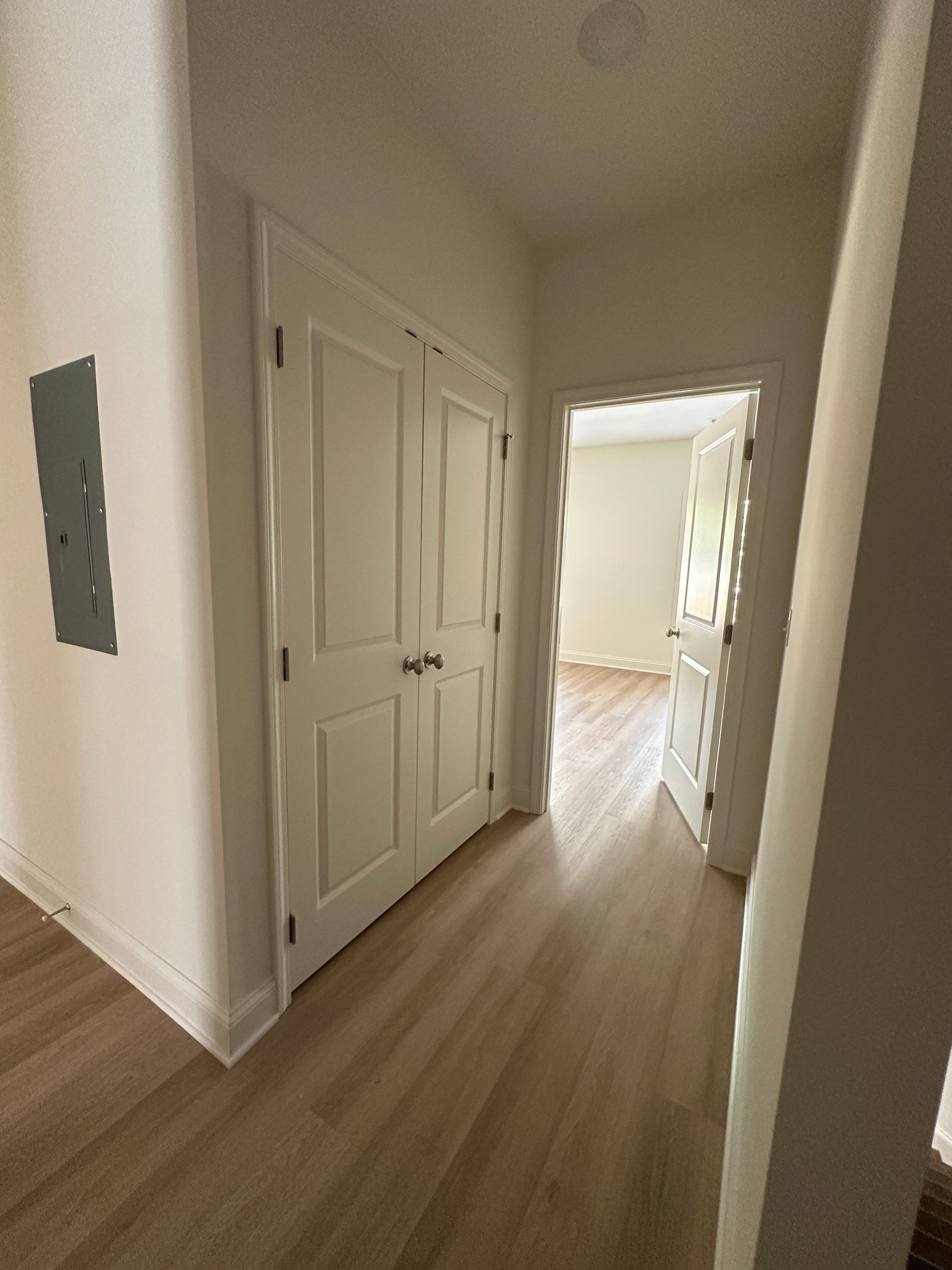 Narrow hallway with white doors, light wood floors, and bright window light at the far end.