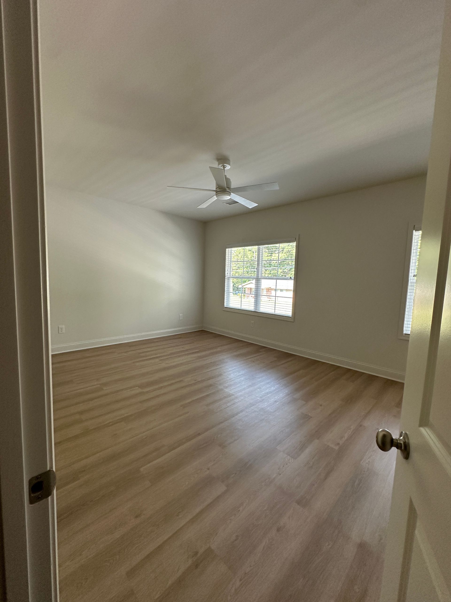 Empty beige bedroom with wood floors, white ceiling fan, and two windows.