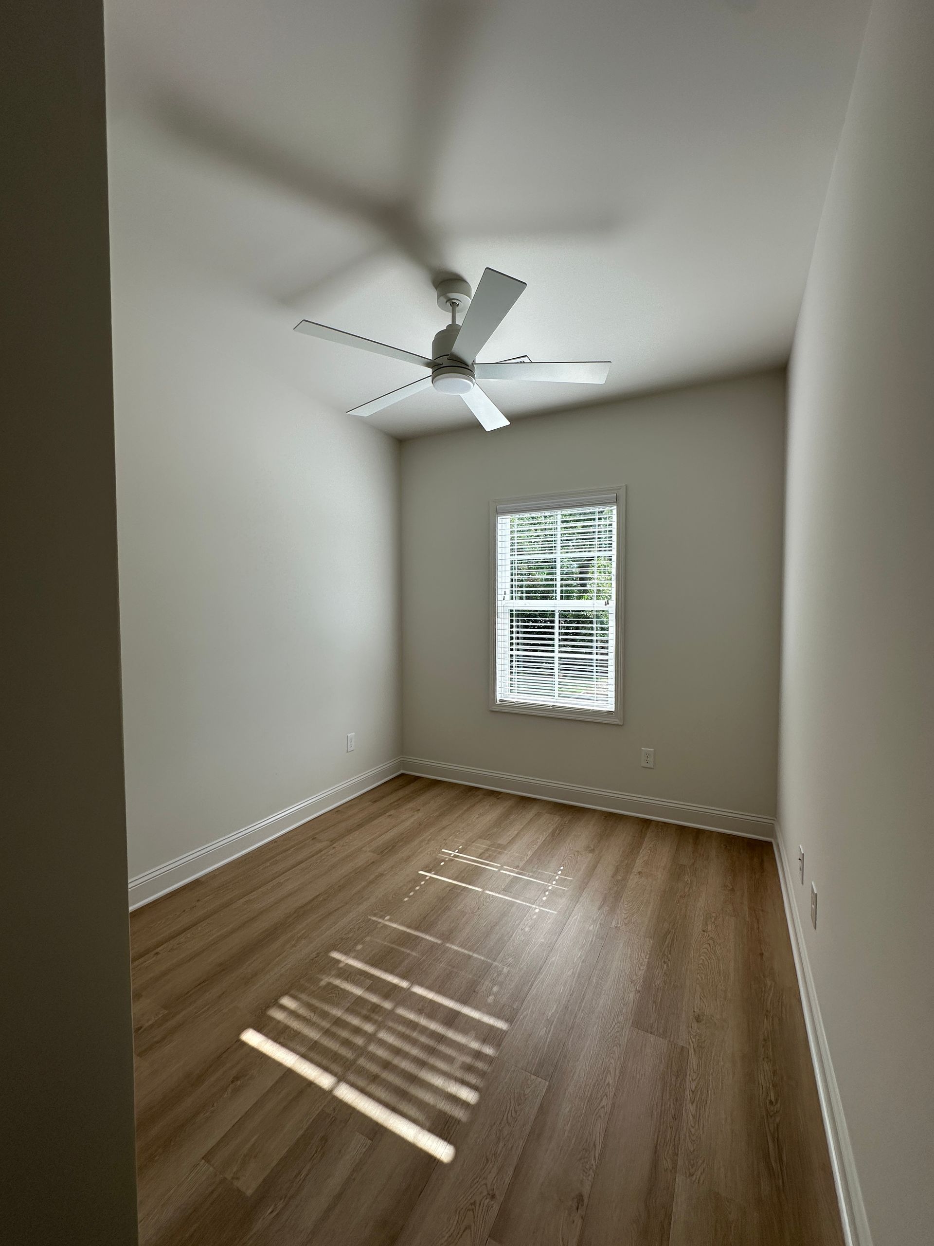 Empty white bedroom with wood floor, window blinds, and a ceiling fan with light.