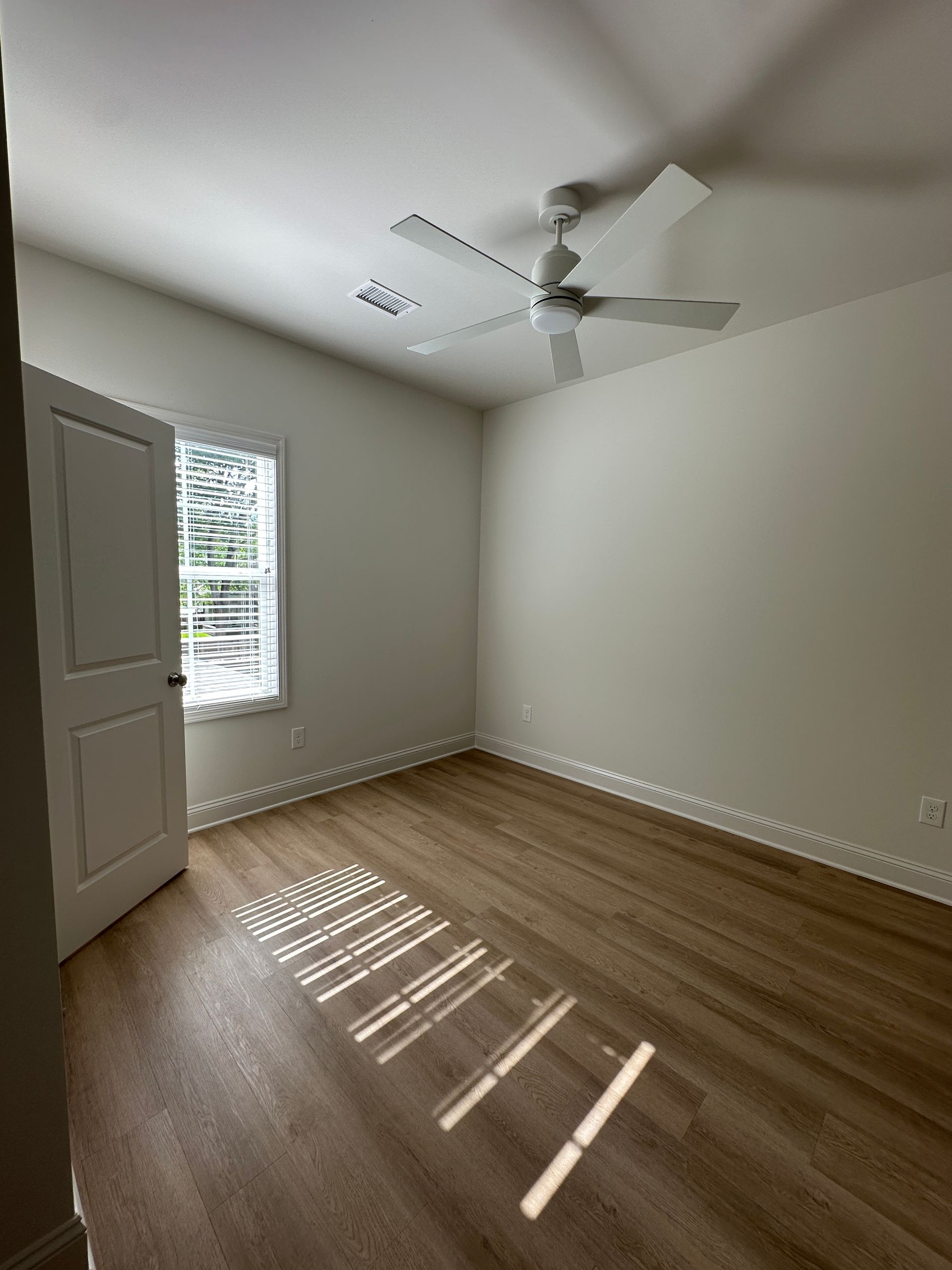 Empty bedroom with white walls, wood floor, window blinds, and a ceiling fan.