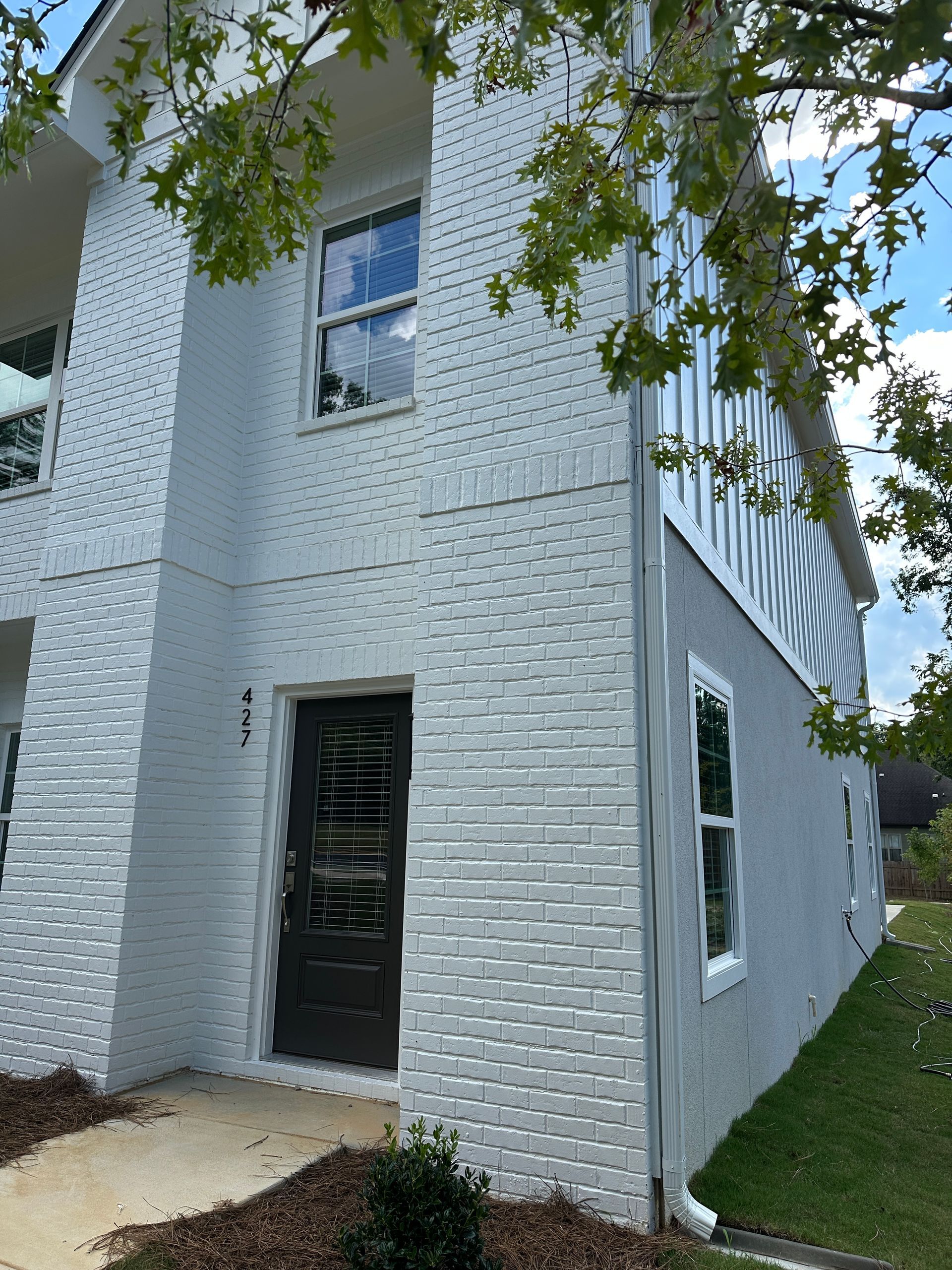 White brick two-story house exterior with black door, windows, and tree branches in the foreground