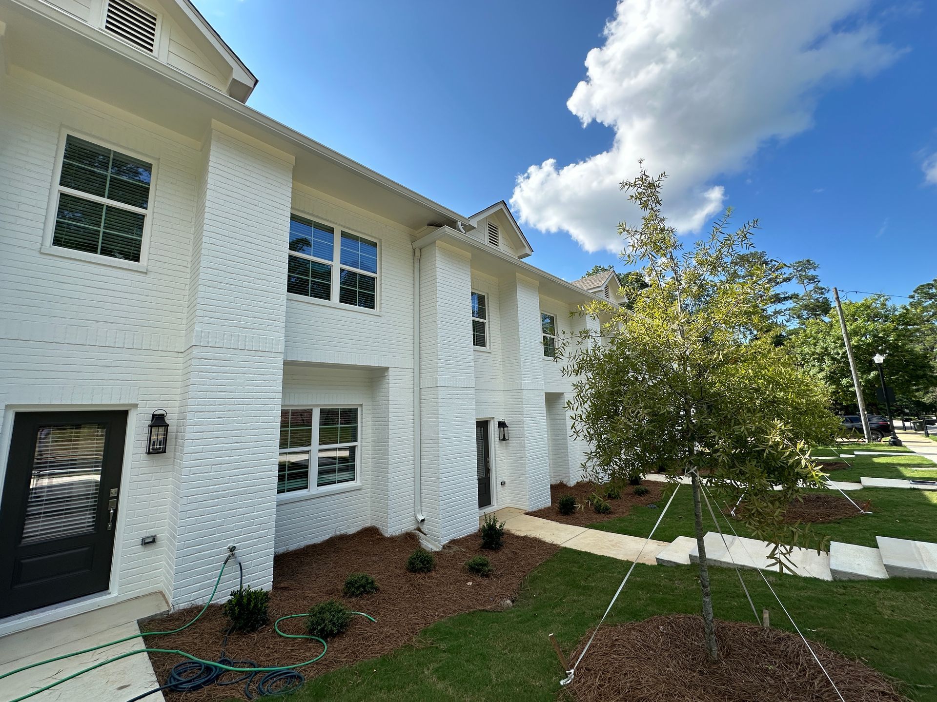 White apartment building with black doors and windows, a walkway, green lawn, and a tree under a blue sky.