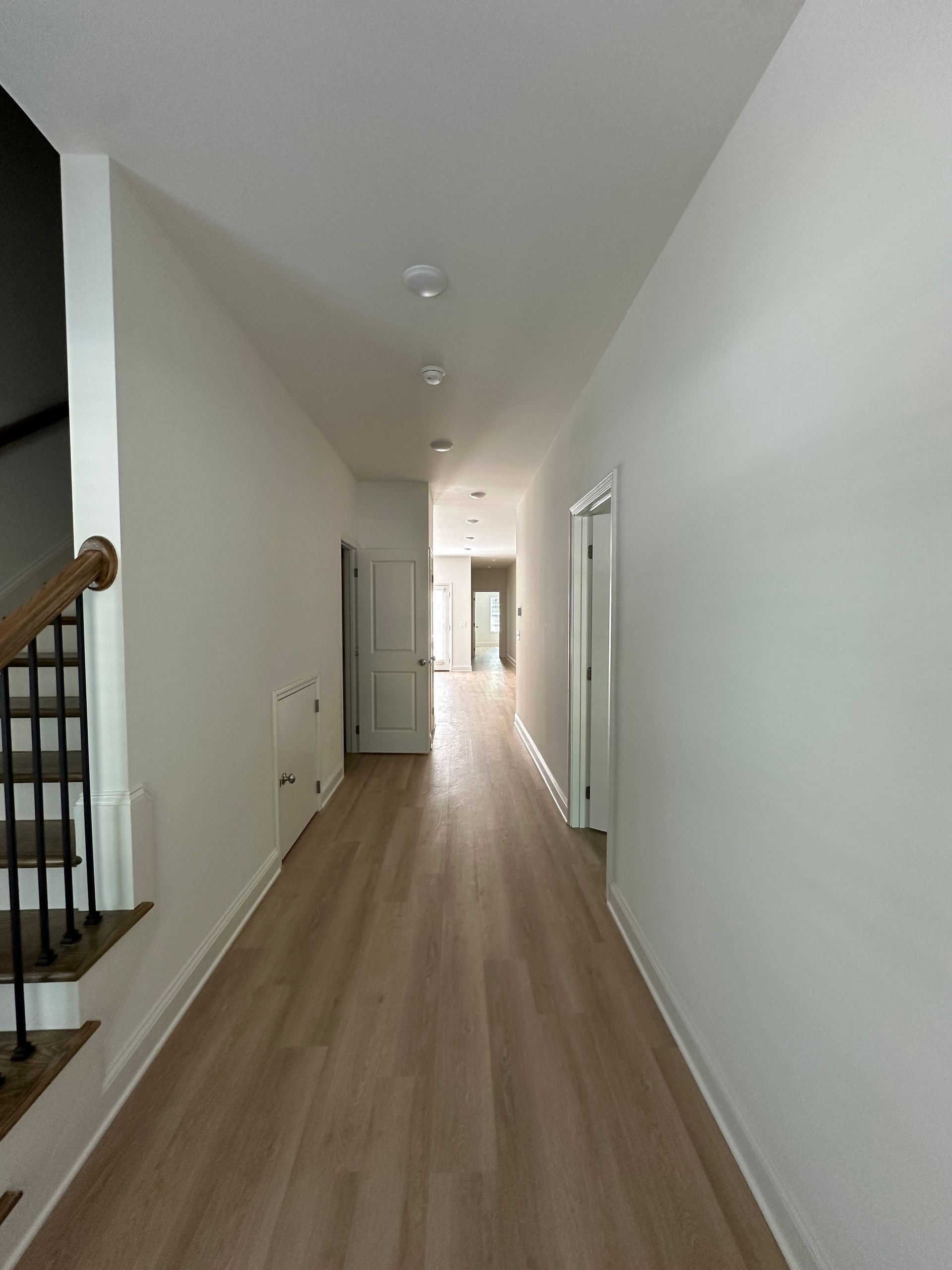 Long, narrow hallway with light wood floors, white walls, and stairs visible on the left.