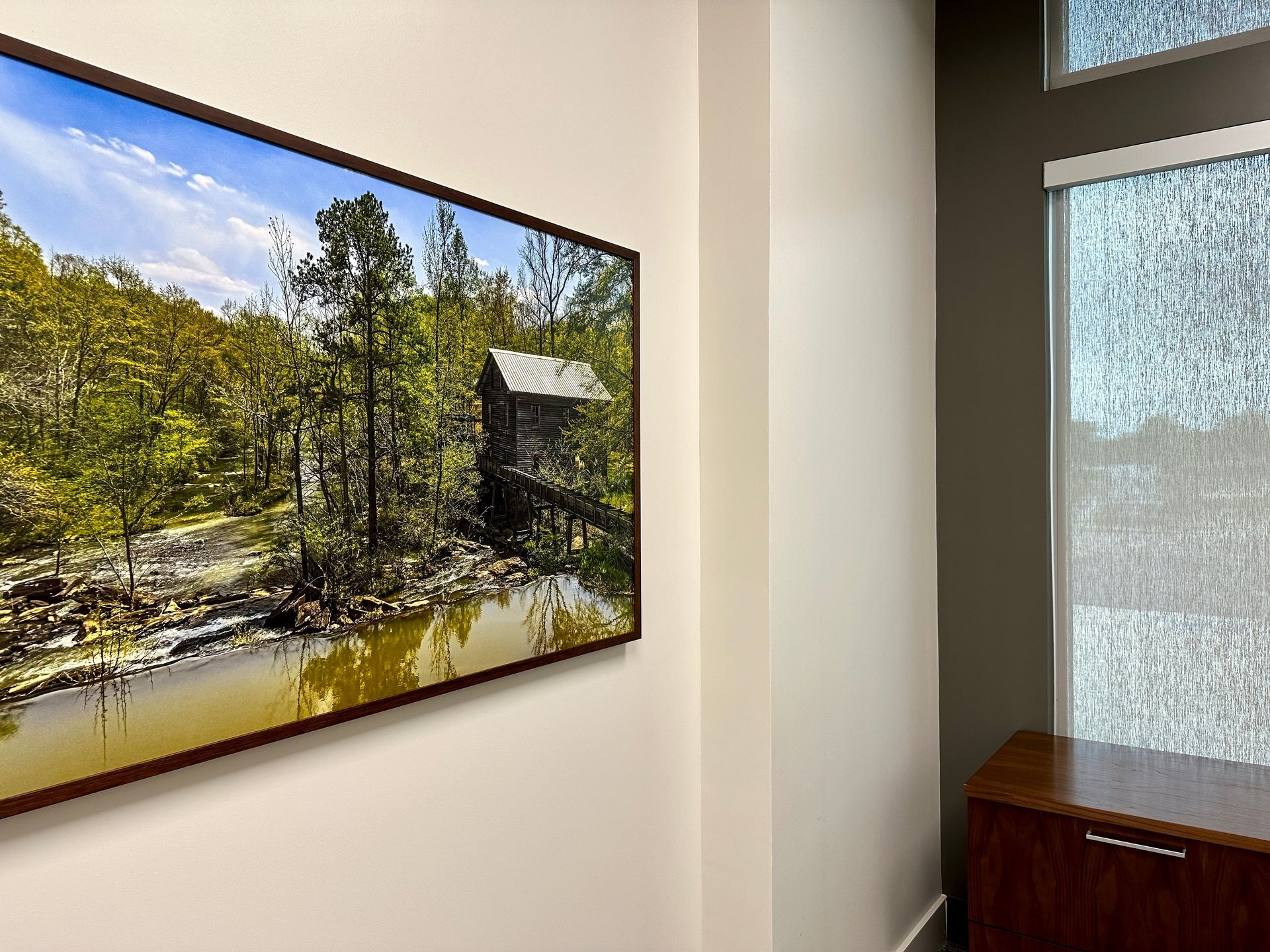 A picture of a cabin in the woods is hanging on a wall next to a window.