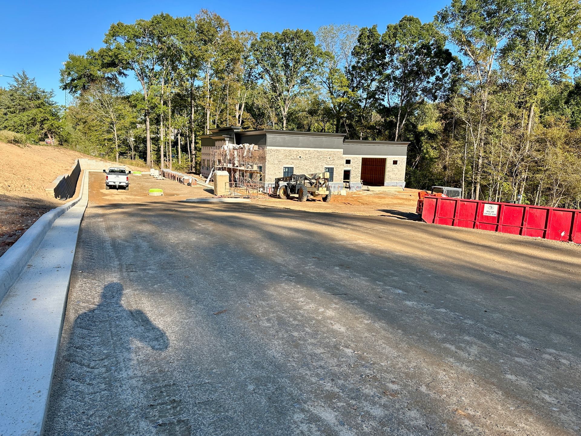 Construction site with a partially built stone building, gravel road, and trees under a blue sky.