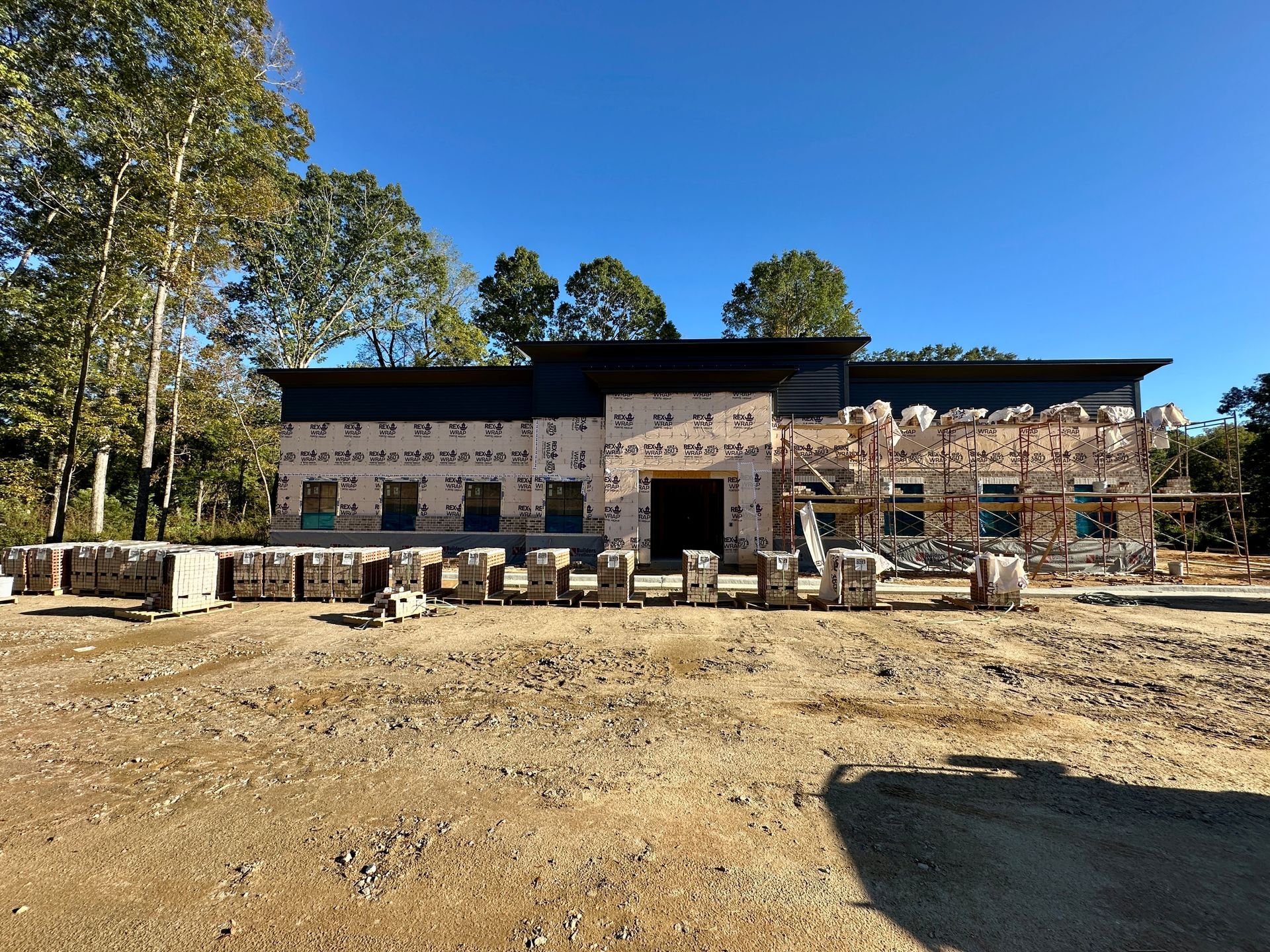 Building under construction; exterior view, beige siding, black roof, sunny day, surrounded by trees.