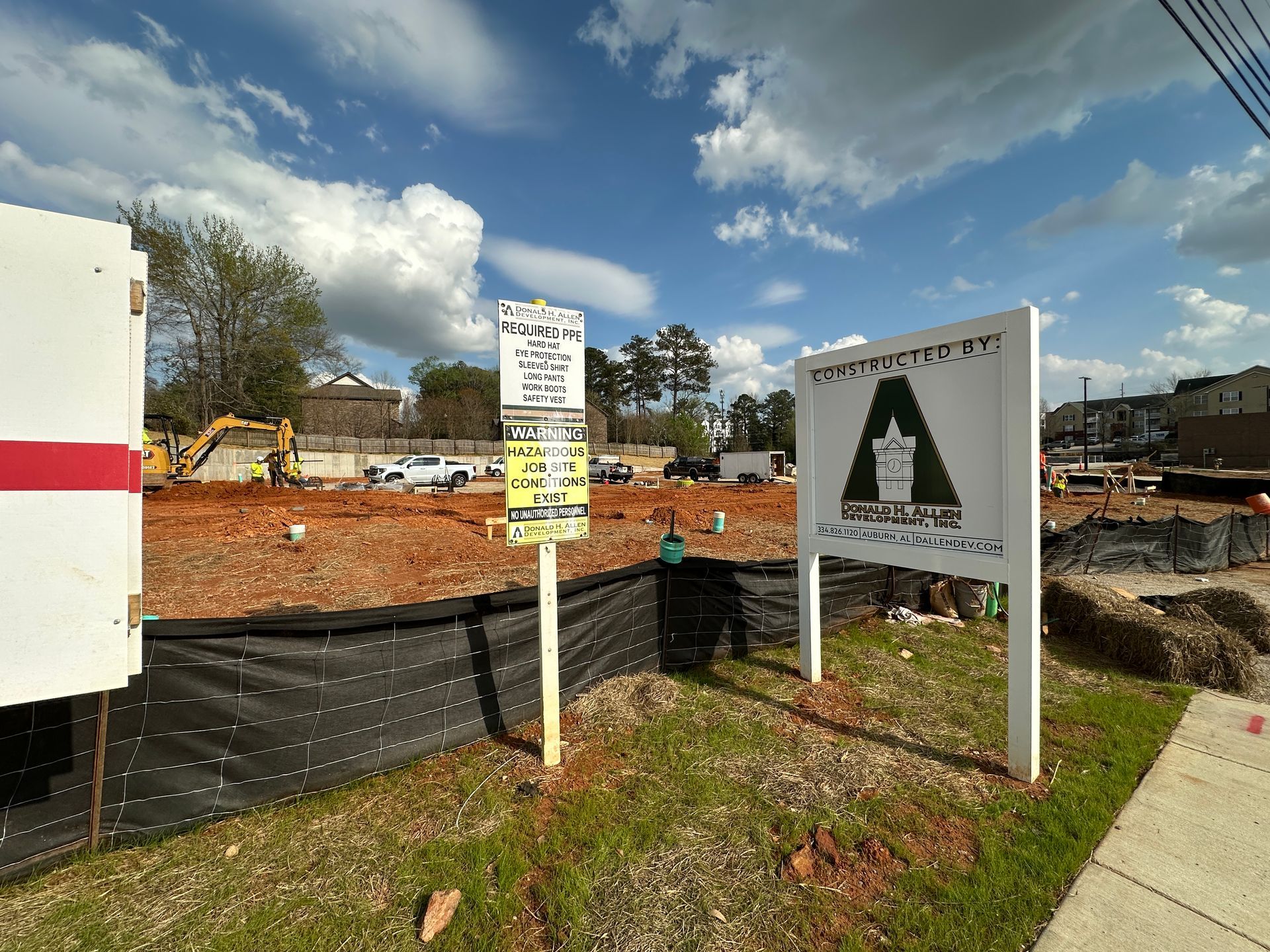 A construction site with two signs, a yellow excavator, and a black silt fence against a blue, cloudy sky.