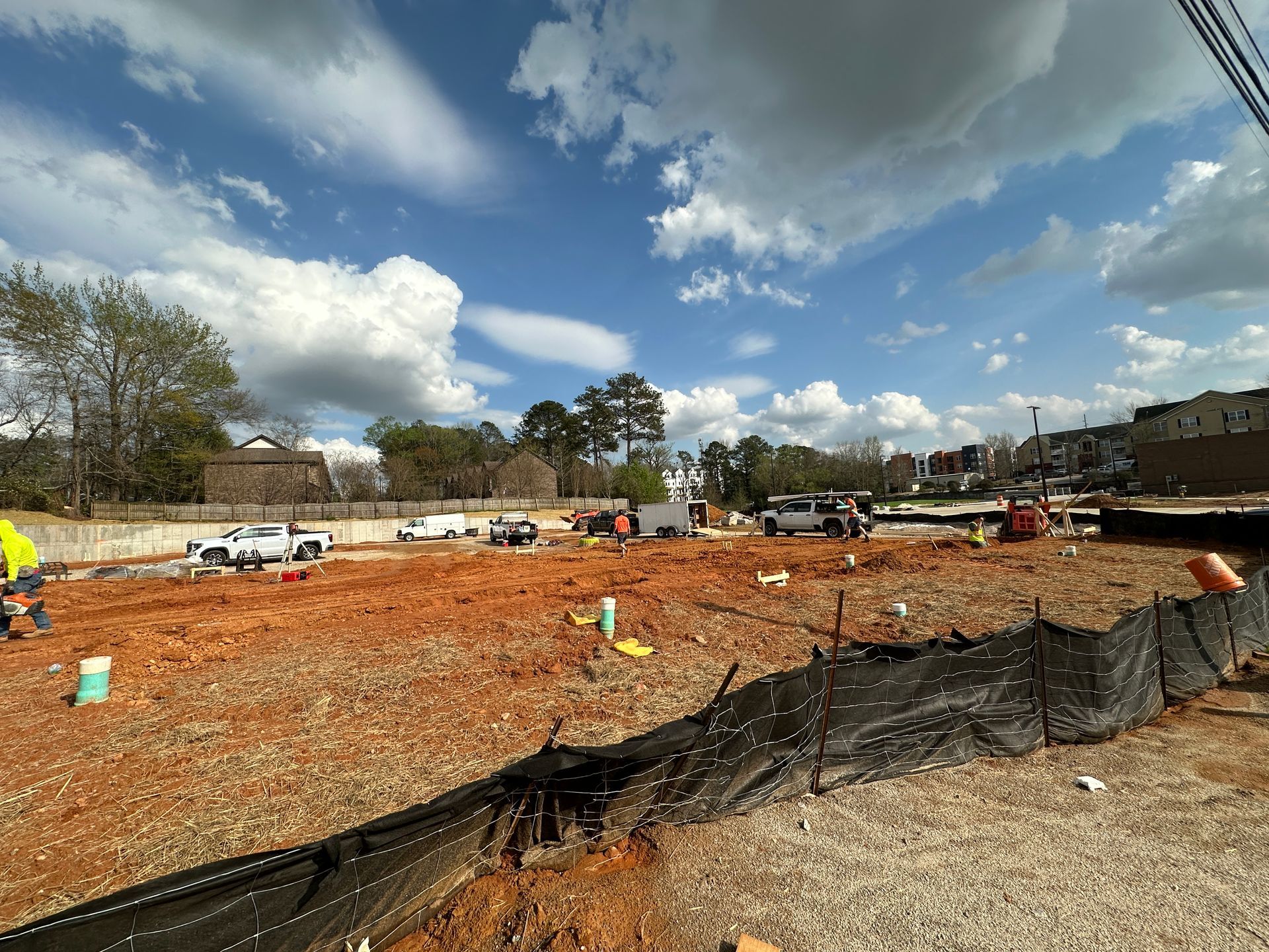 Construction crew members work on a large, orange dirt lot enclosed by a black silt fence under a cloudy blue sky.