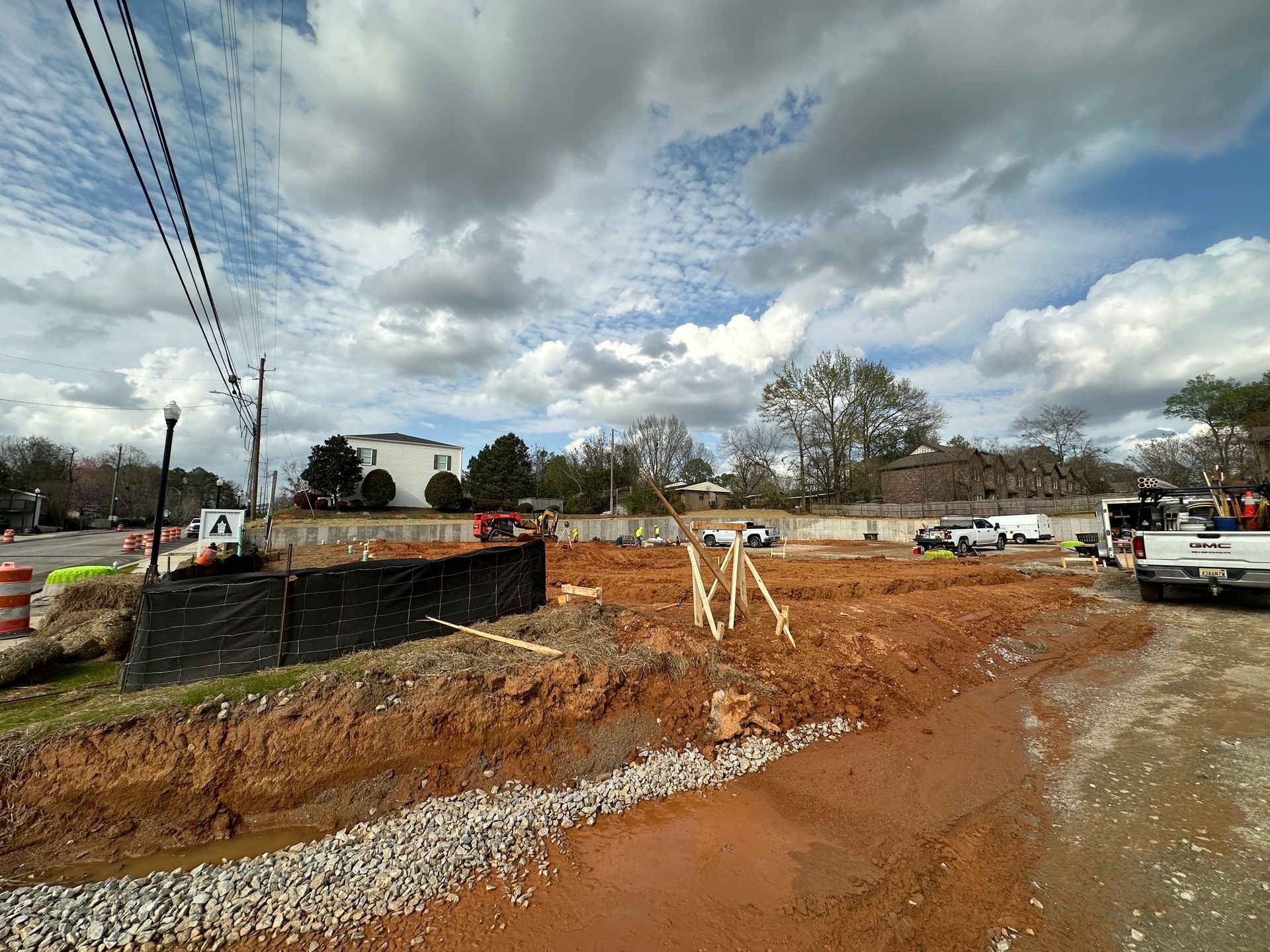 A construction site with exposed red earth, a gravel border, and a black silt fence under a cloudy sky.