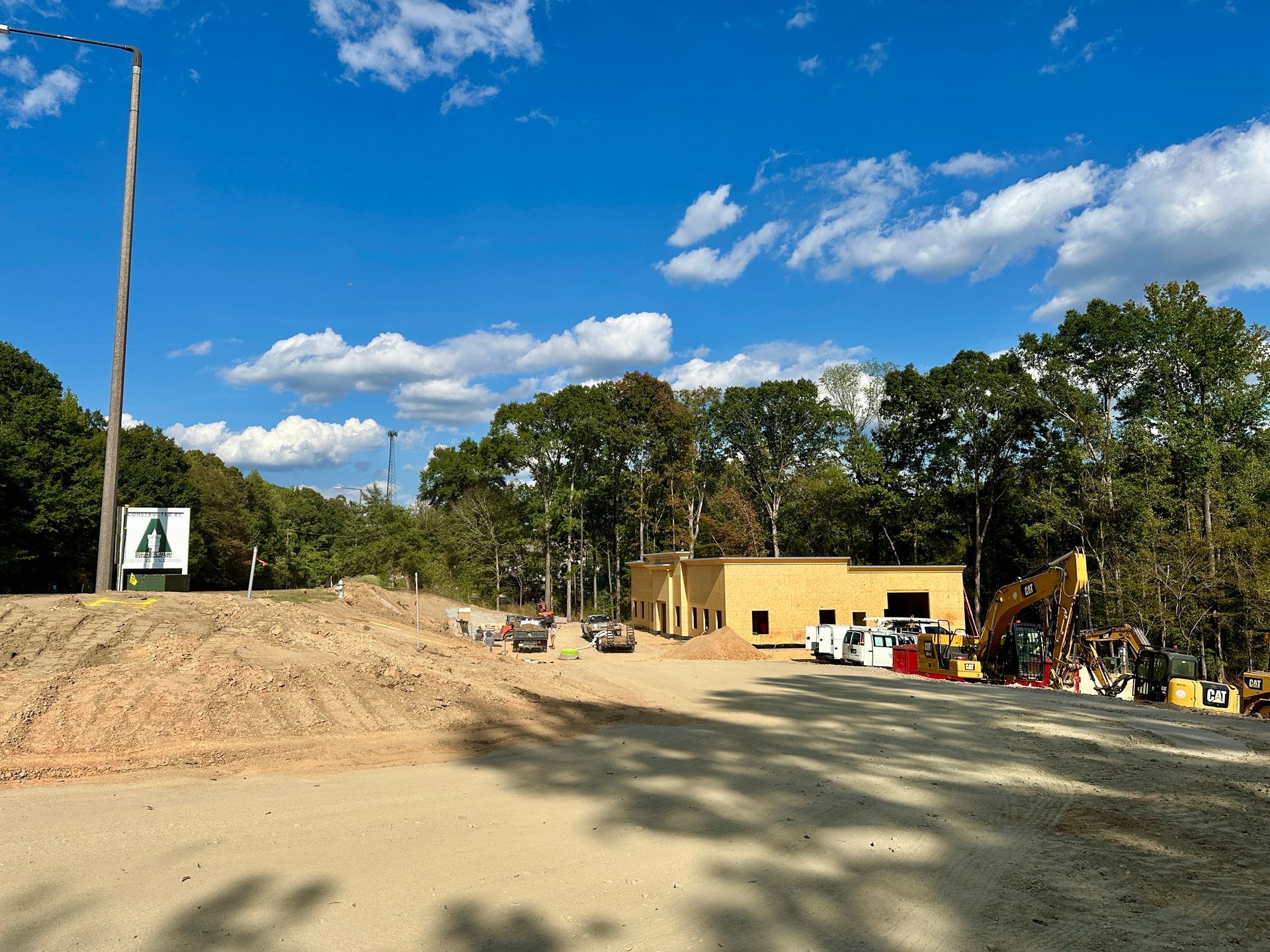 Construction site with building, equipment, dirt piles, and trees under a blue sky with clouds.
