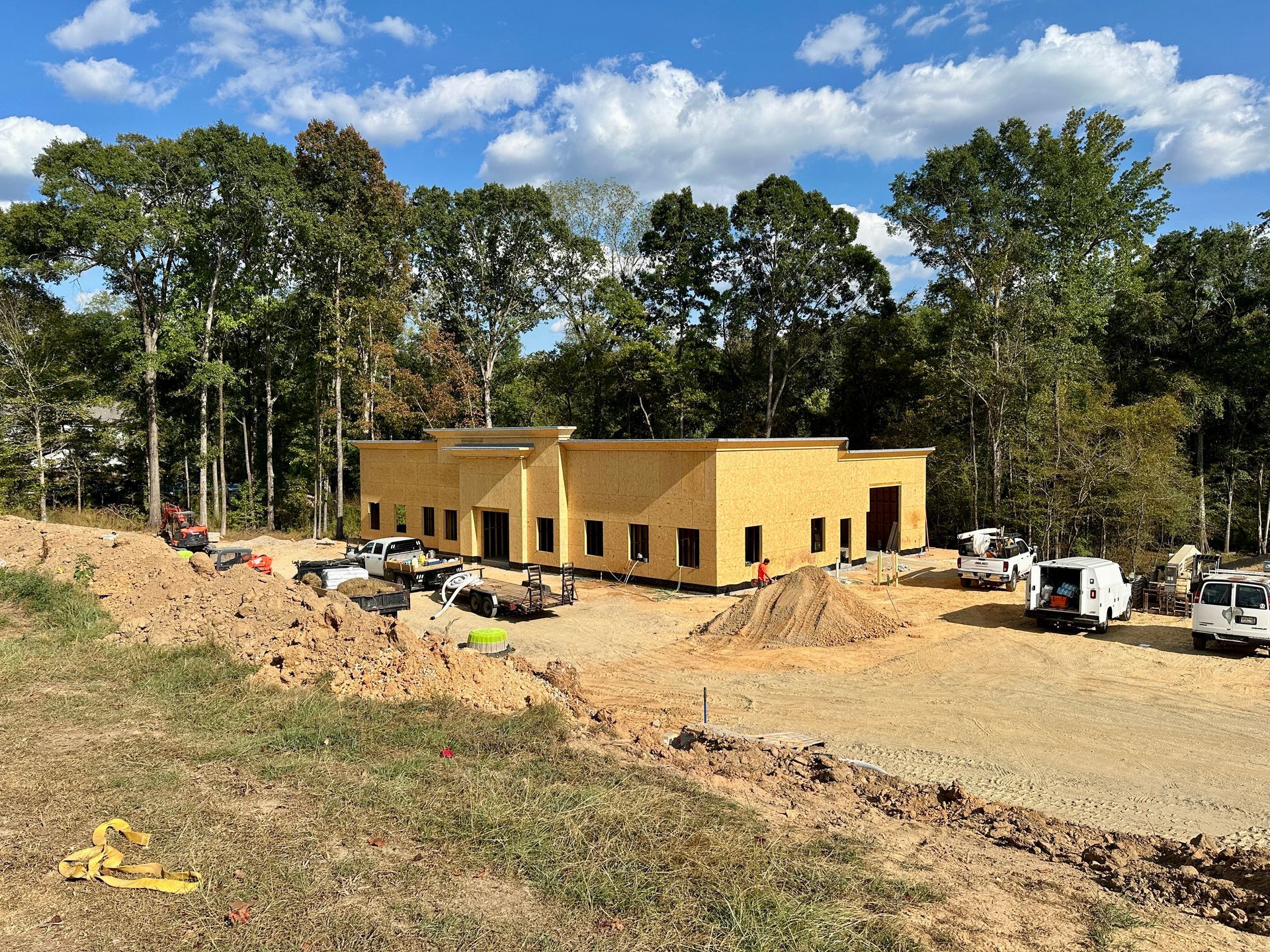 Construction site with a wood-framed building surrounded by dirt, trucks, and trees under a blue sky.