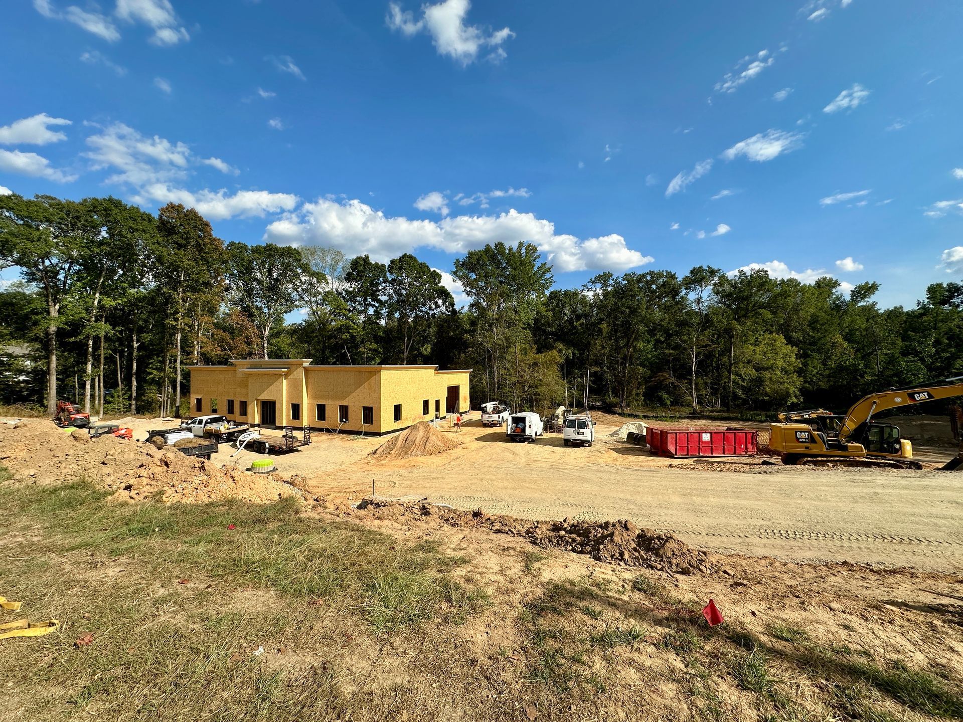Construction site with a building frame, earth moving equipment, and trees under a blue sky.