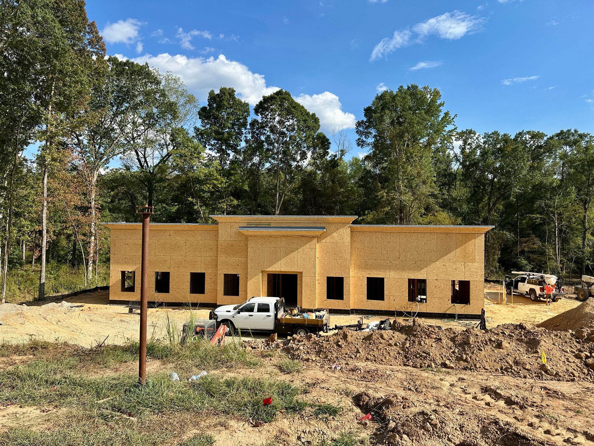 Building under construction: wood-framed structure with a pickup truck in front and a tree-lined backdrop on a sunny day.