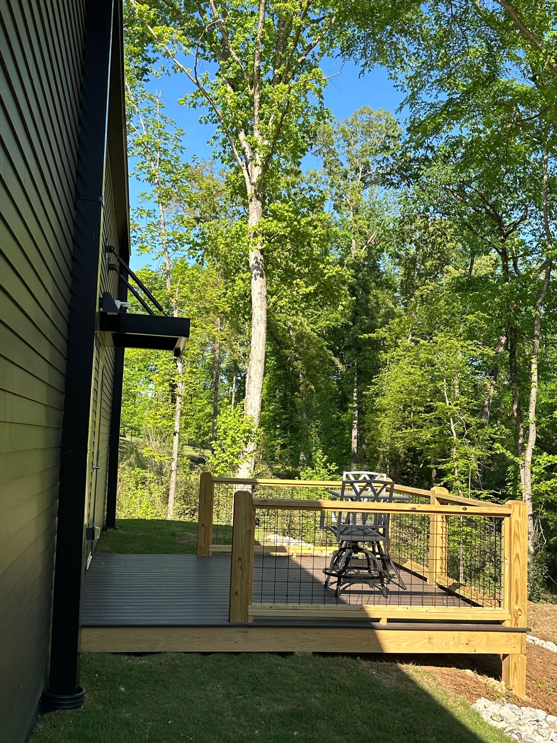 A dark wood deck with a metal railing and patio furniture overlooks a sunlit forest of green trees.