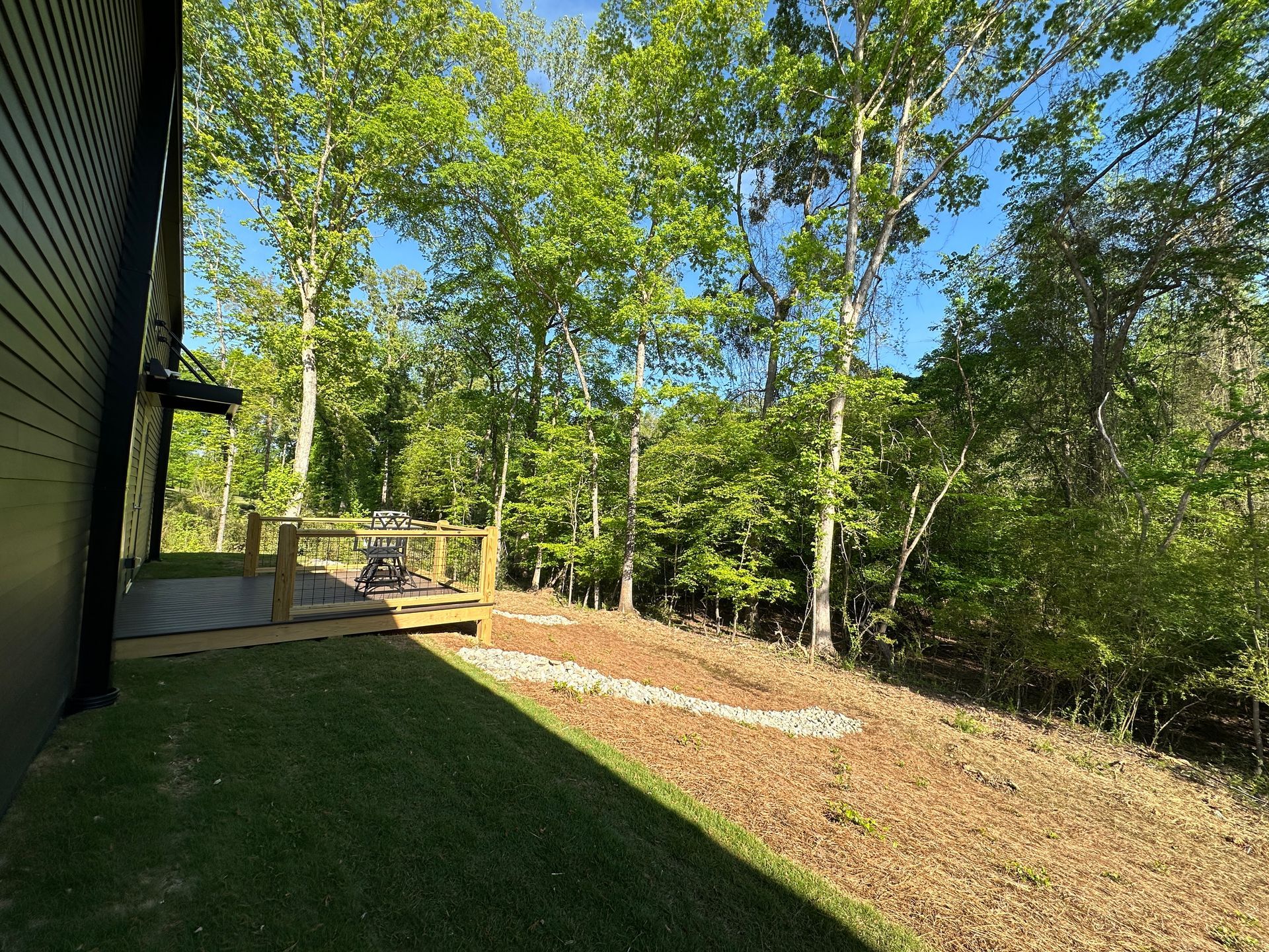 A wooden deck sits next to a dark house wall, overlooking a grassy lawn and a wooded area on a sunny day.