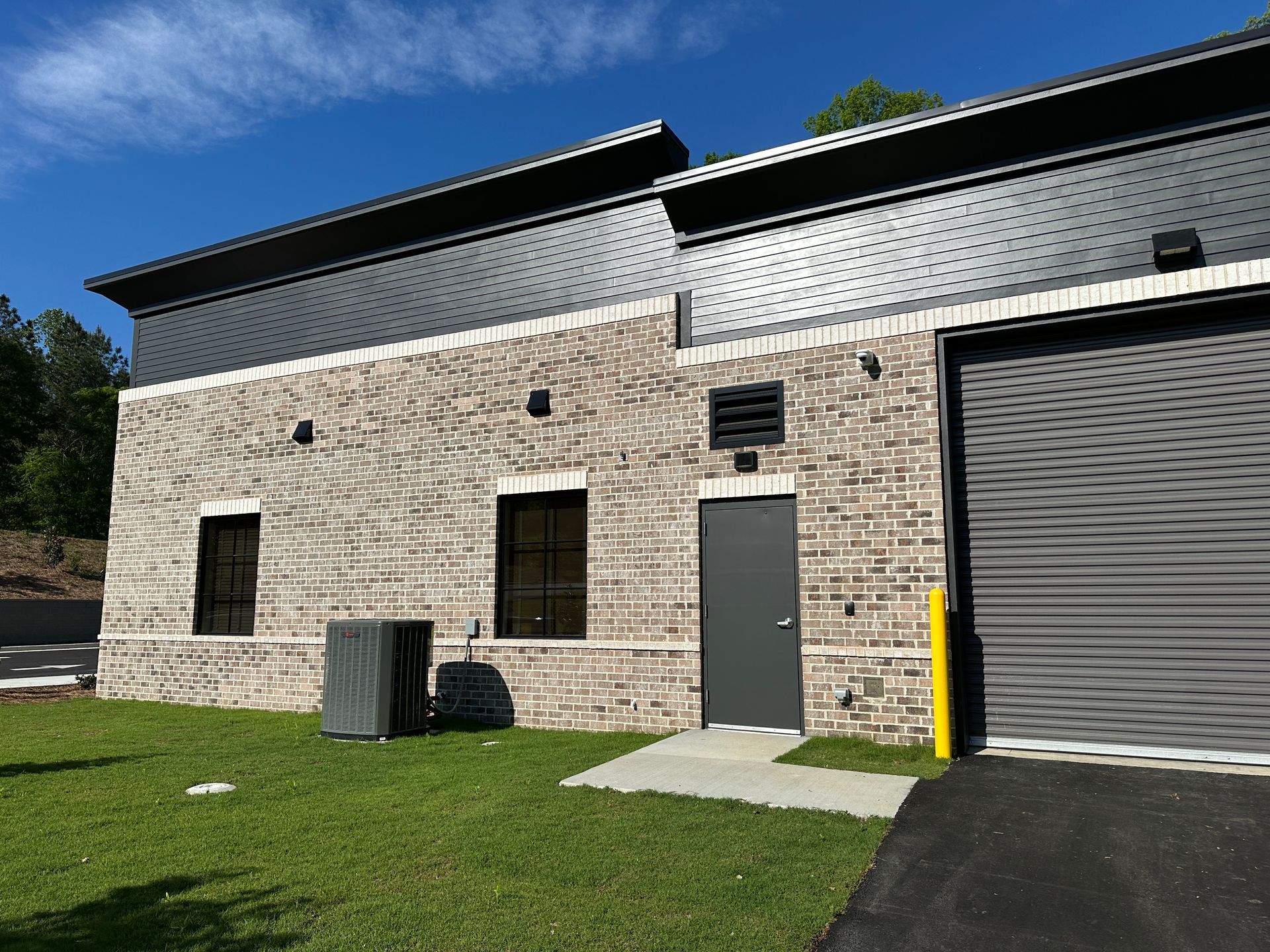 A modern, light-colored brick warehouse building featuring a grey metal door, two windows, and a large roll-up garage door.