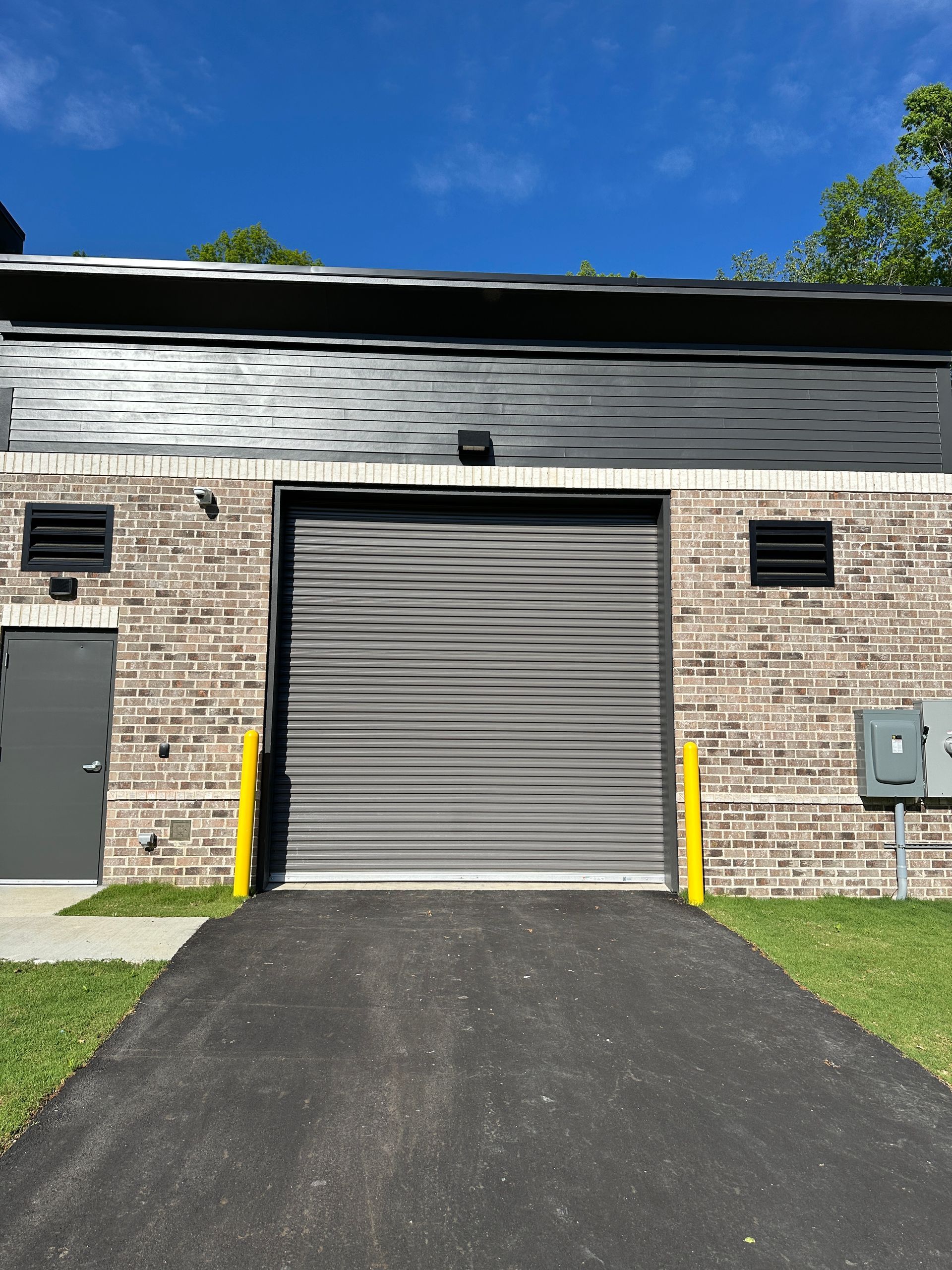 A brick commercial building with a gray roll-up metal door and a gray side entry door under a clear blue sky.