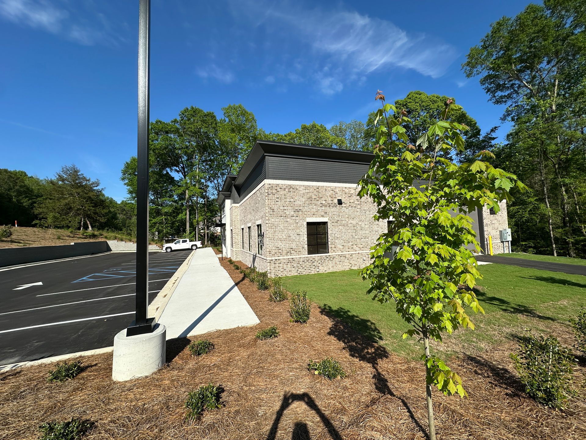 A stone building with a dark roof next to a paved parking lot under a bright blue sky, with a small tree in the foreground.
