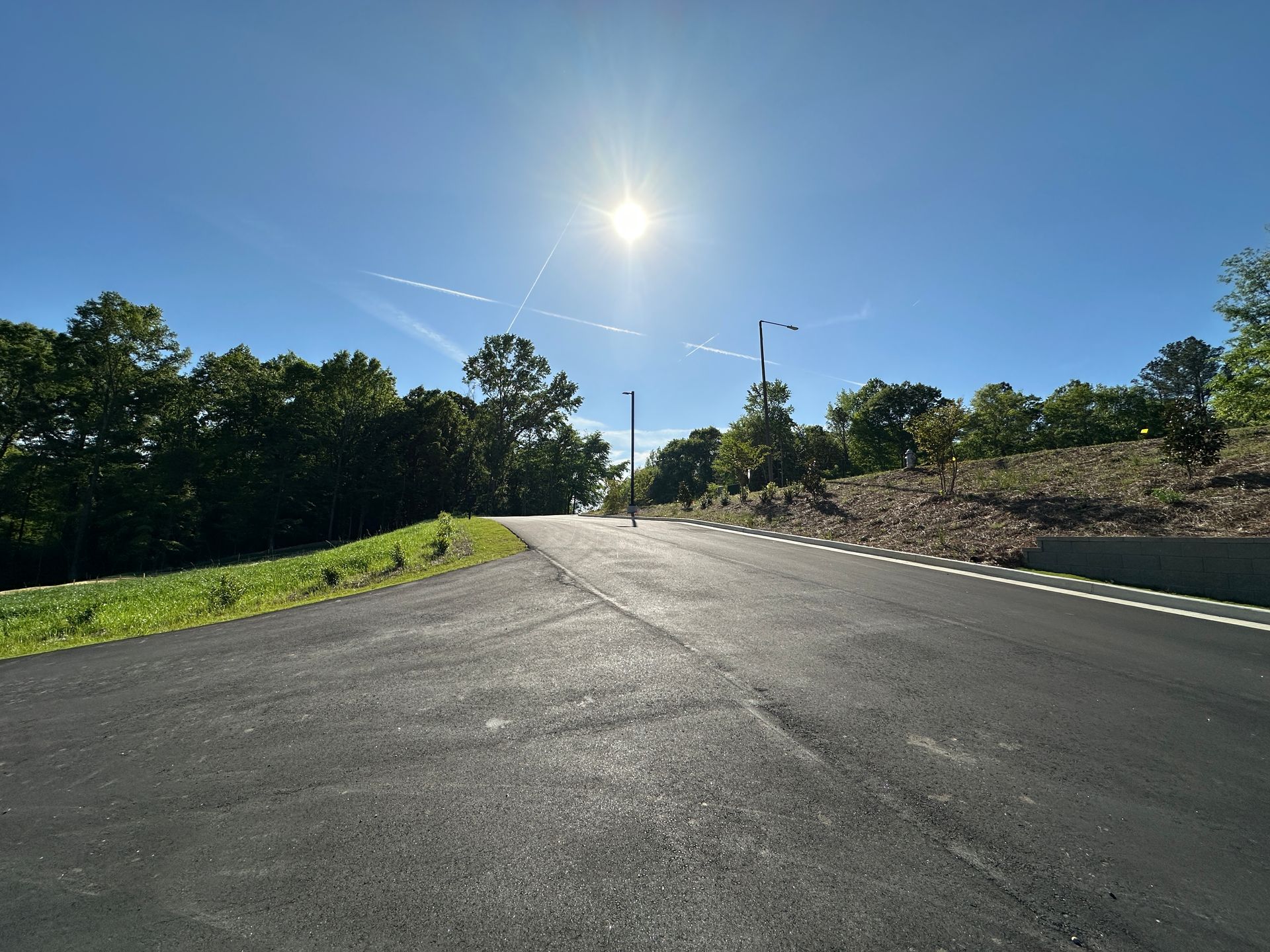 A sunlit, paved road stretches forward, flanked by grassy areas and woods under a clear blue sky.