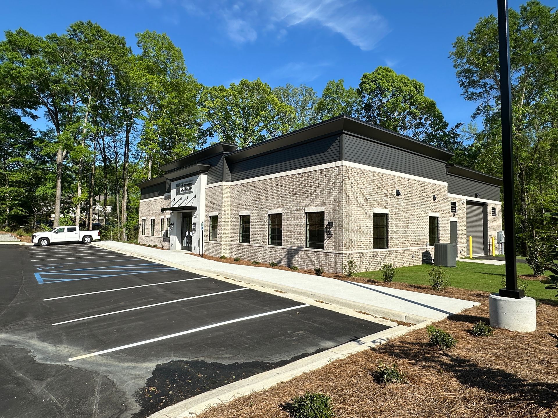 A single-story commercial building with light gray textured brick and a dark metal roof, next to a paved parking lot.