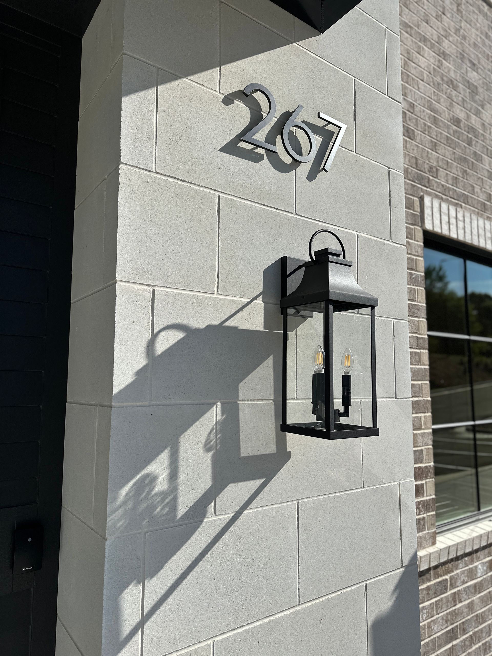 The number 267 in black metal digits mounted on a light gray concrete block wall beside a black outdoor lantern.