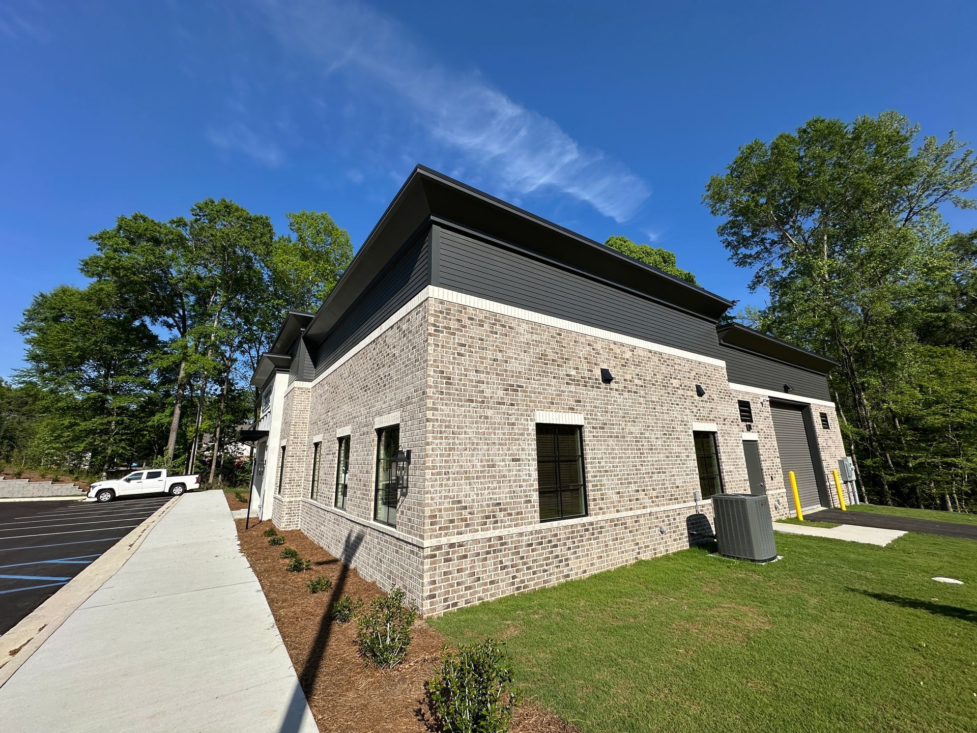 A modern commercial building with a light gray brick exterior, a dark metal roof, and a sidewalk against a blue sky.