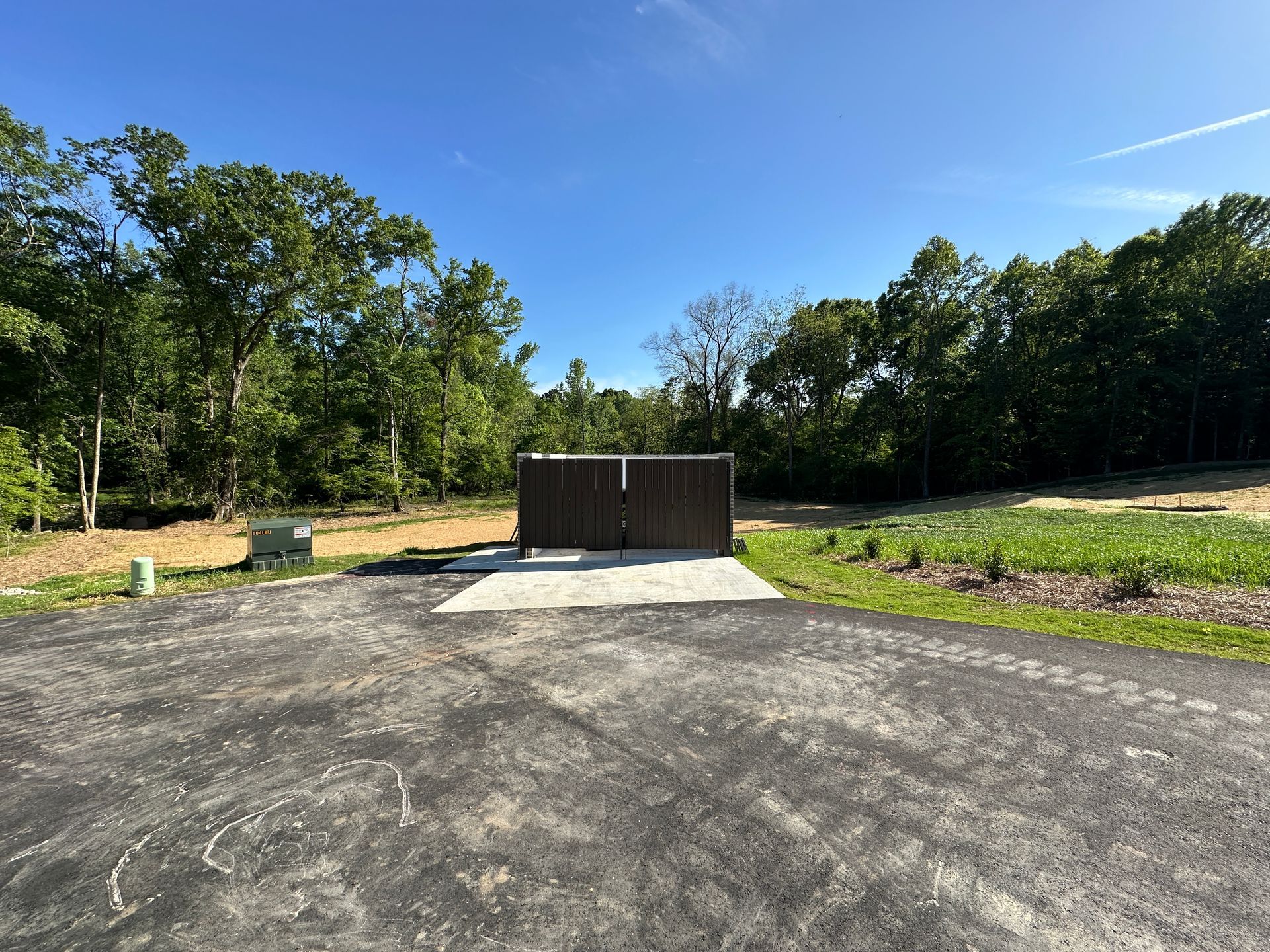A paved driveway leads to a concrete pad with a dark brown metal trash enclosure, surrounded by trees under a blue sky.
