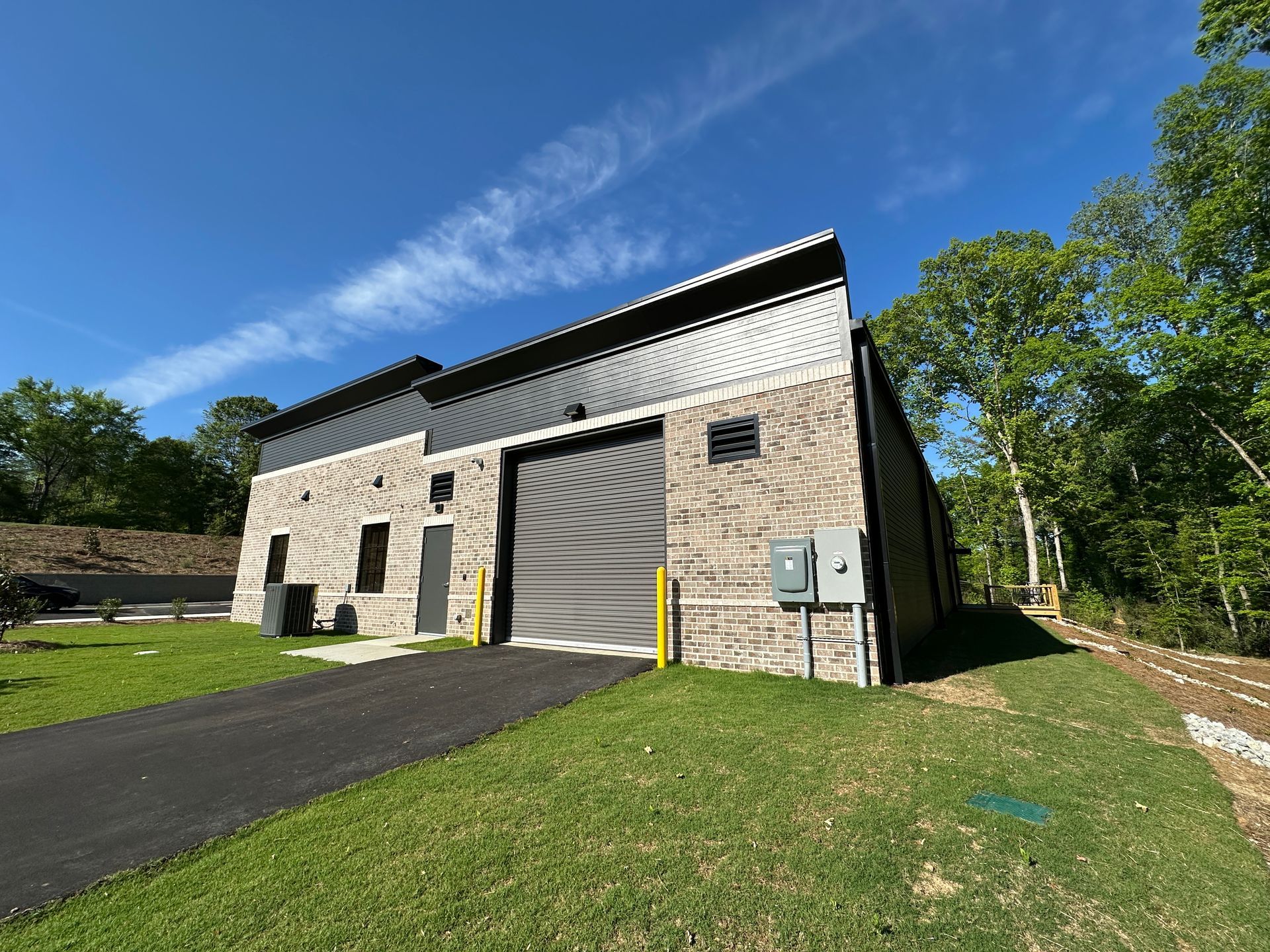 A modern, tan brick industrial building with a large rolling door and dark metal roof under a bright blue sky.