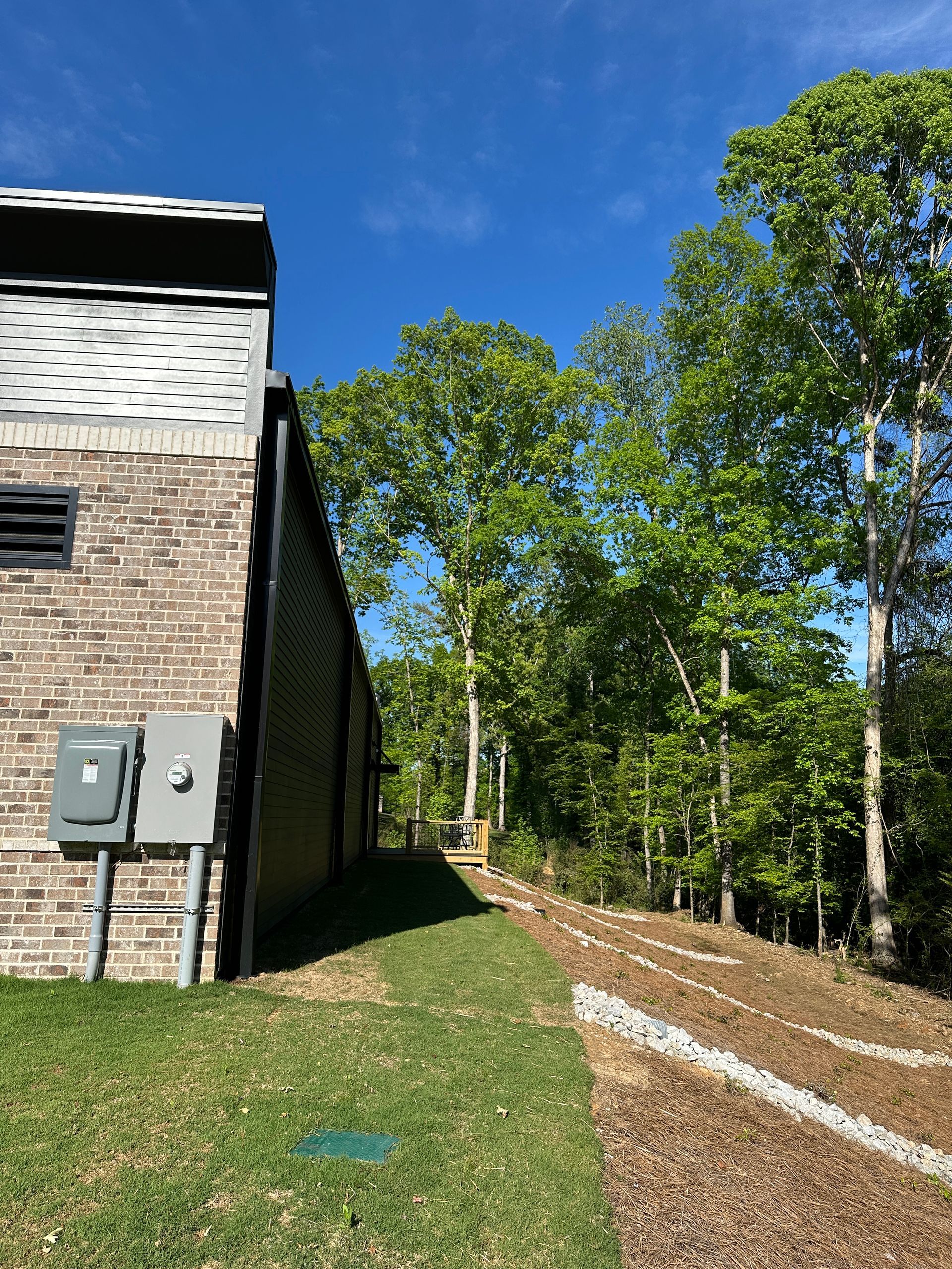A brick building side wall next to a grassy path sloping down toward a wooded area under a bright blue sky.