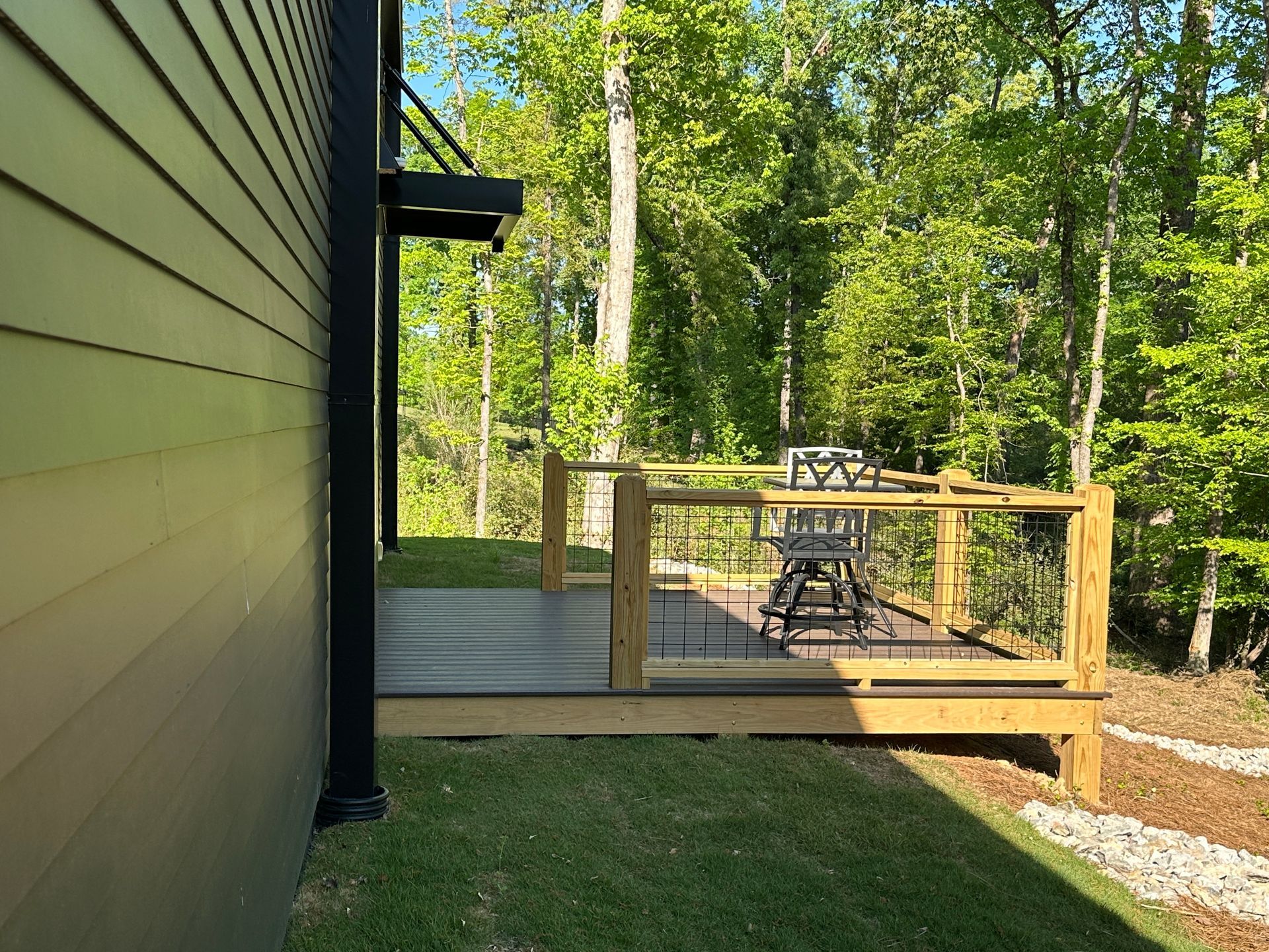 A wooden deck with metal railing sits against a house siding, overlooking a lush green forest on a sunny day.