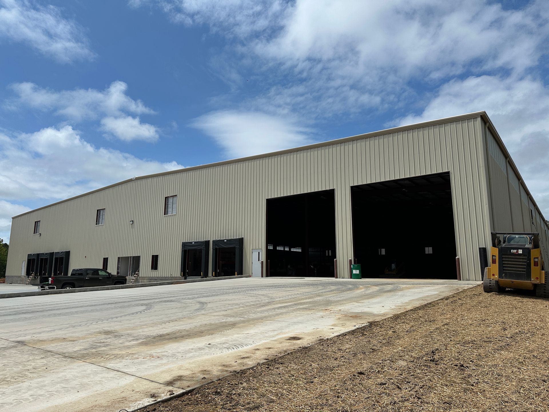 A large, light-colored industrial building with two open bays; a small construction vehicle is parked on the right.