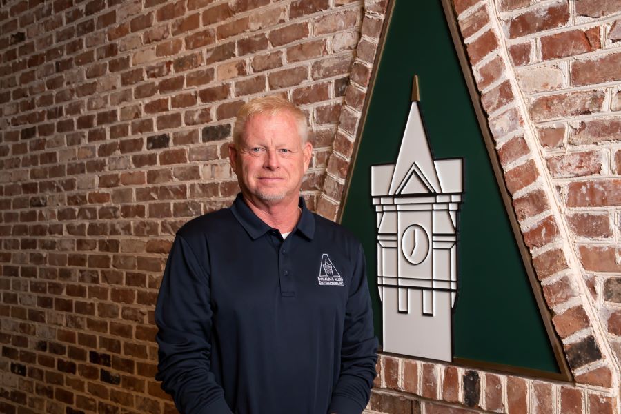 A man in a blue shirt is standing in front of a brick wall.