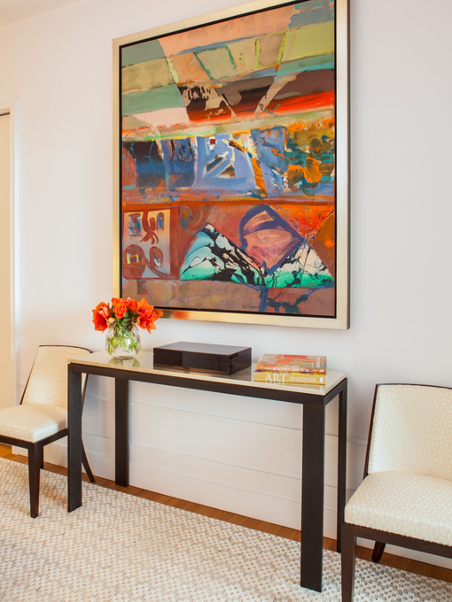 Abstract art above console table with flowers, books, electronics, flanked by two chairs on a white rug.